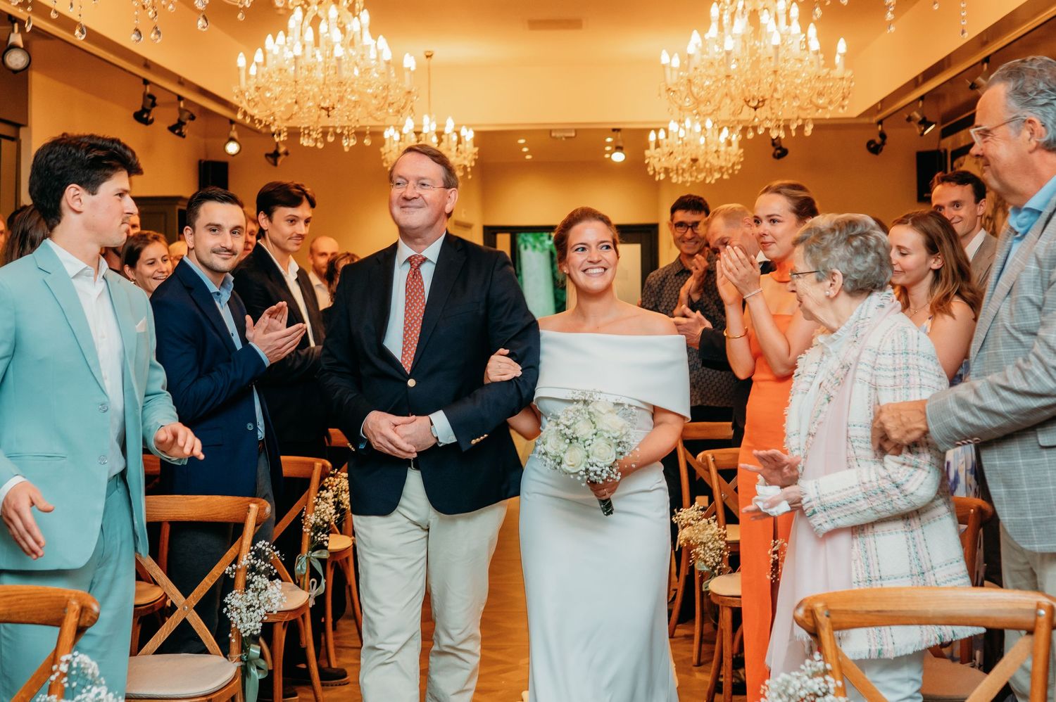 Bride in off-shoulder gown walks the aisle with escort amid applauding guests in elegant chandelier venue.