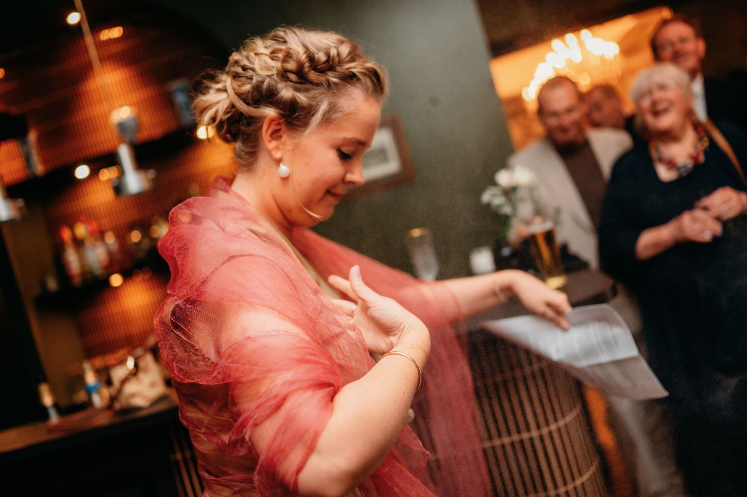 Woman in a pink sheer dress laughs joyfully while guests celebrate in the background at a wedding reception.