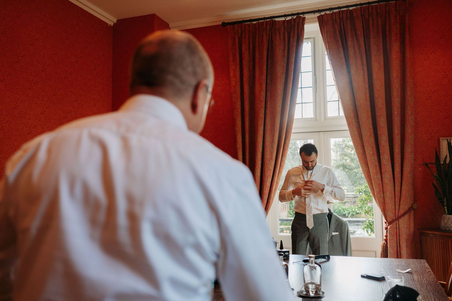 Groom in white shirt stands in a red-walled room, watching another man adjust his tie by a tall window with red curtains.