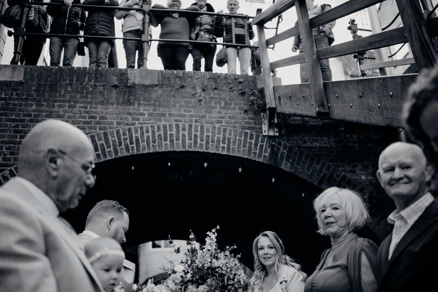 Bruiloftsgasten op brug kijken naar feest beneden, zwart wit bruidsfotografie in Den Bosch en omgeving Brabant.
