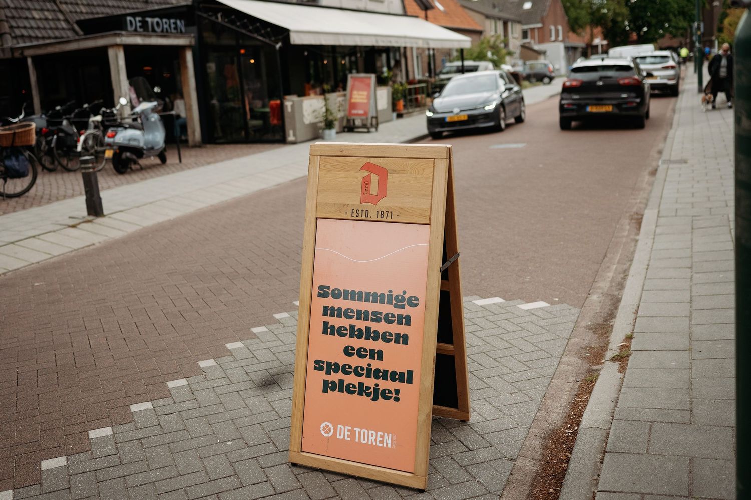 A wooden sandwich board sign with Dutch text outside De Token restaurant on a brick street.
