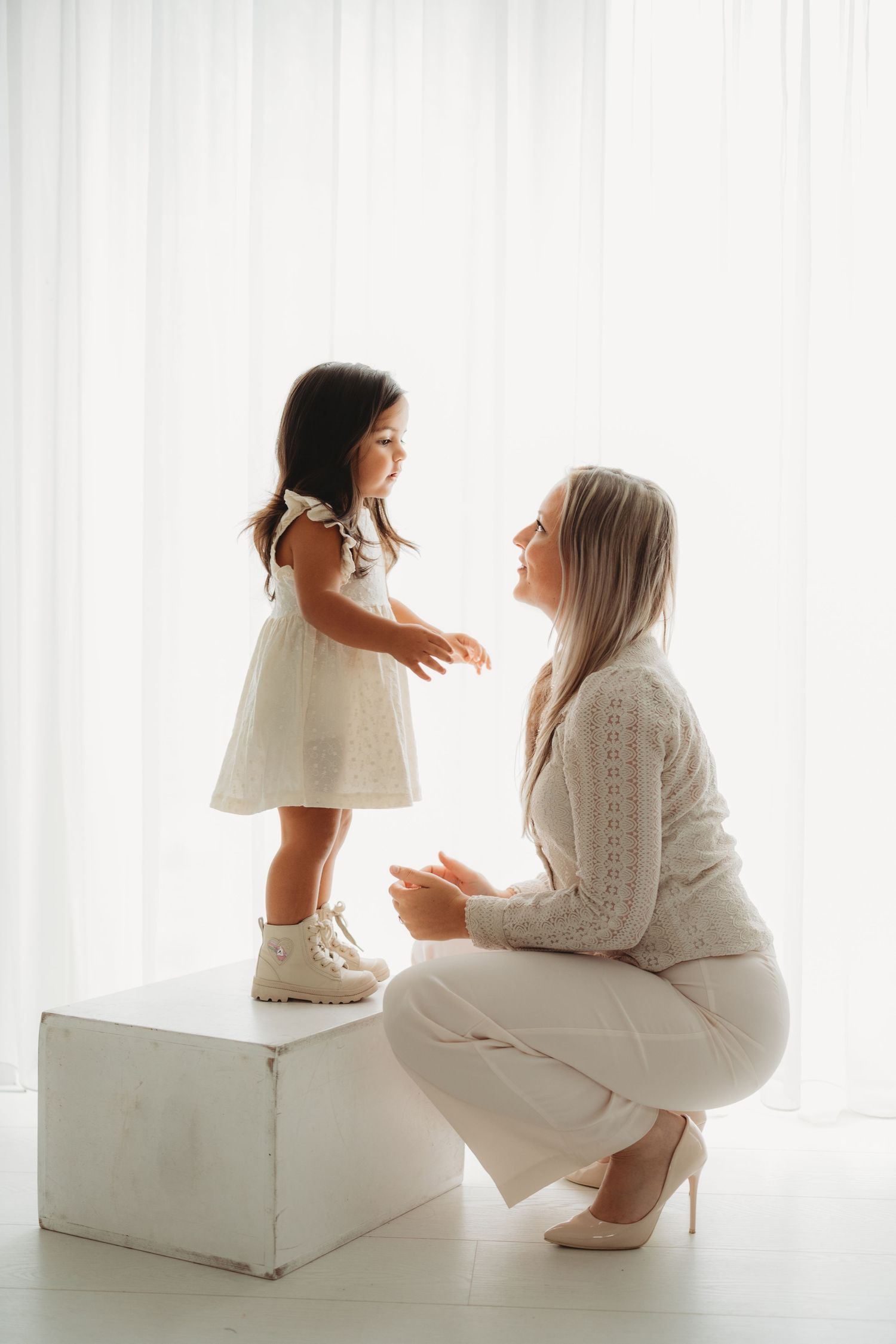 A mother crouches to eye level with her toddler daughter standing on a white cube, both dressed in elegant white outfits.