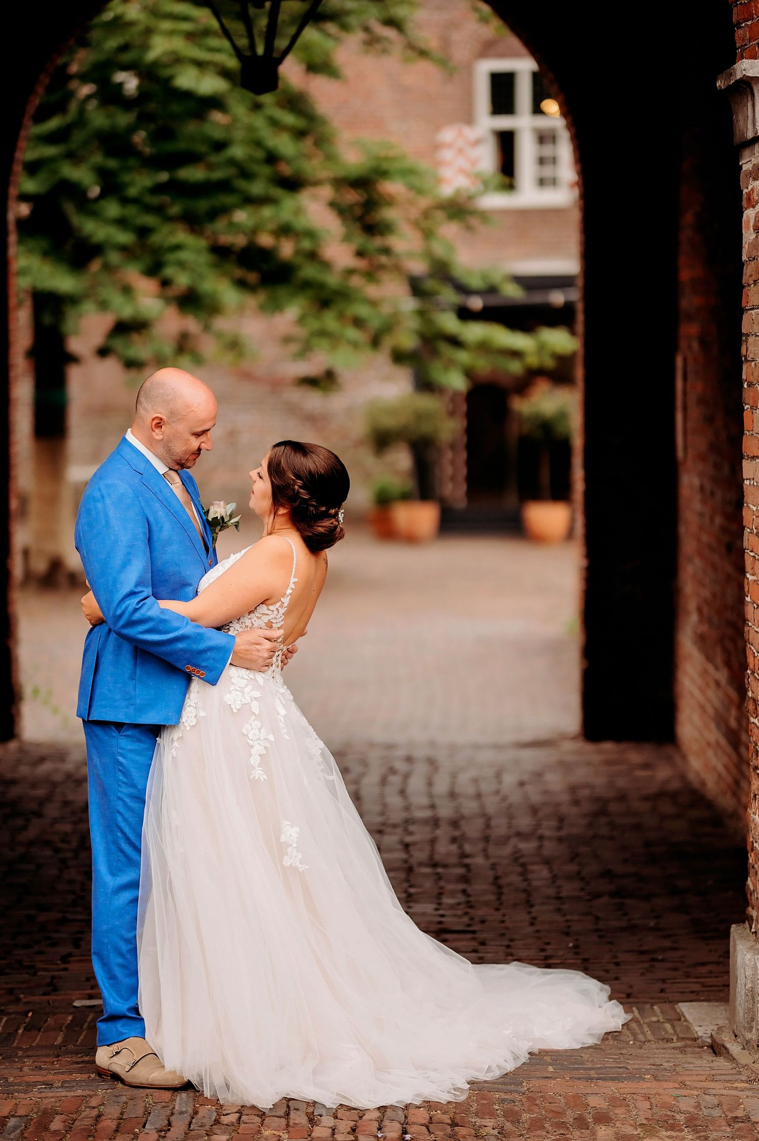 Bride and groom share romantic moment under brick archway in courtyard wearing blue suit and white gown.