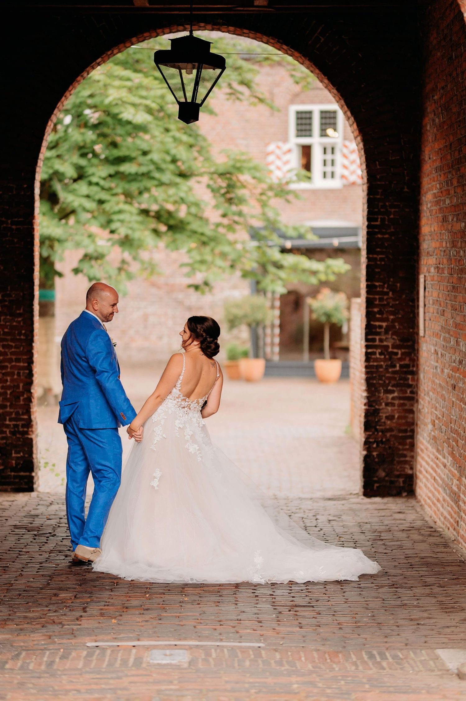 Bride and groom holding hands walking through a brick archway, she in a floral wedding gown, he in blue.