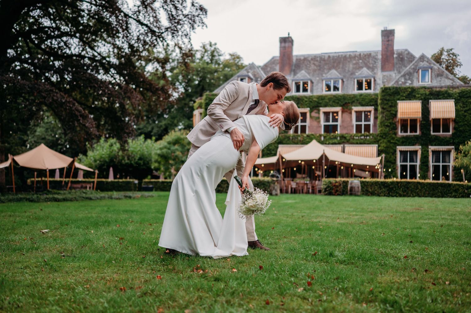 Groom dips bride in romantic kiss on lawn outside grand estate wedding venue.