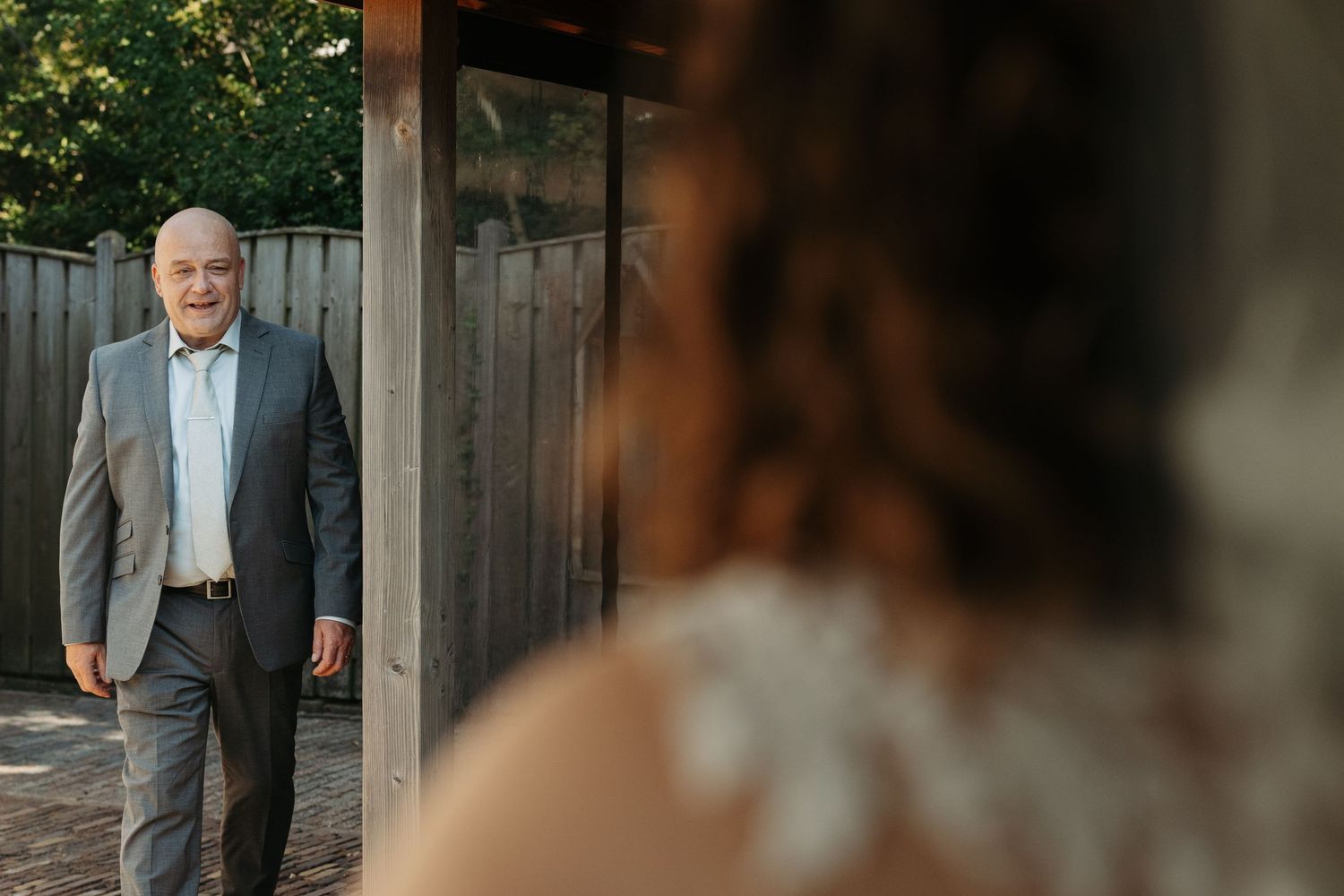 A father in a gray suit smiles warmly as he sees his daughter in a wedding dress for the first time.