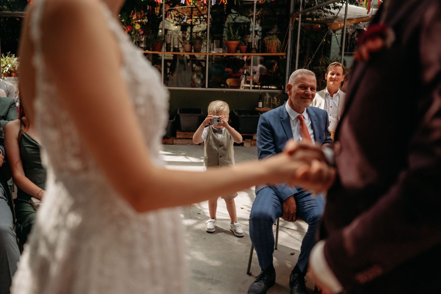 Bruidspaar kijkt elkaar aan tijdens ceremonie onder lichtslingers in kas in Brabant, emotionele trouwfotografie