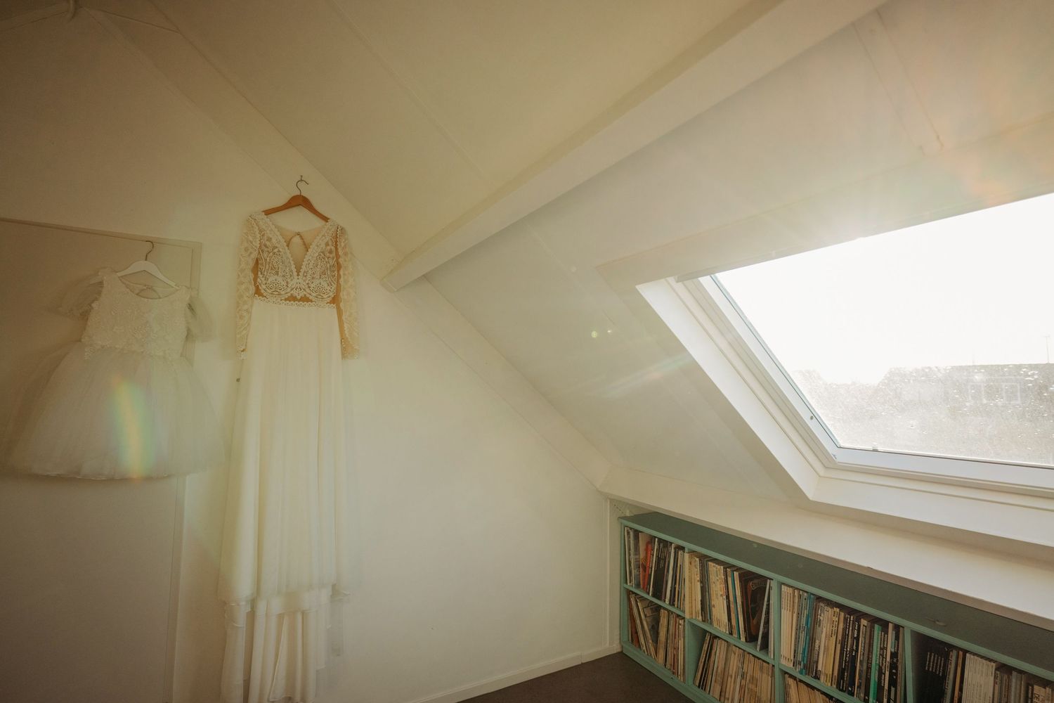 A white wedding dress hangs in a sunlit attic room with built-in bookshelves beneath a skylight window.