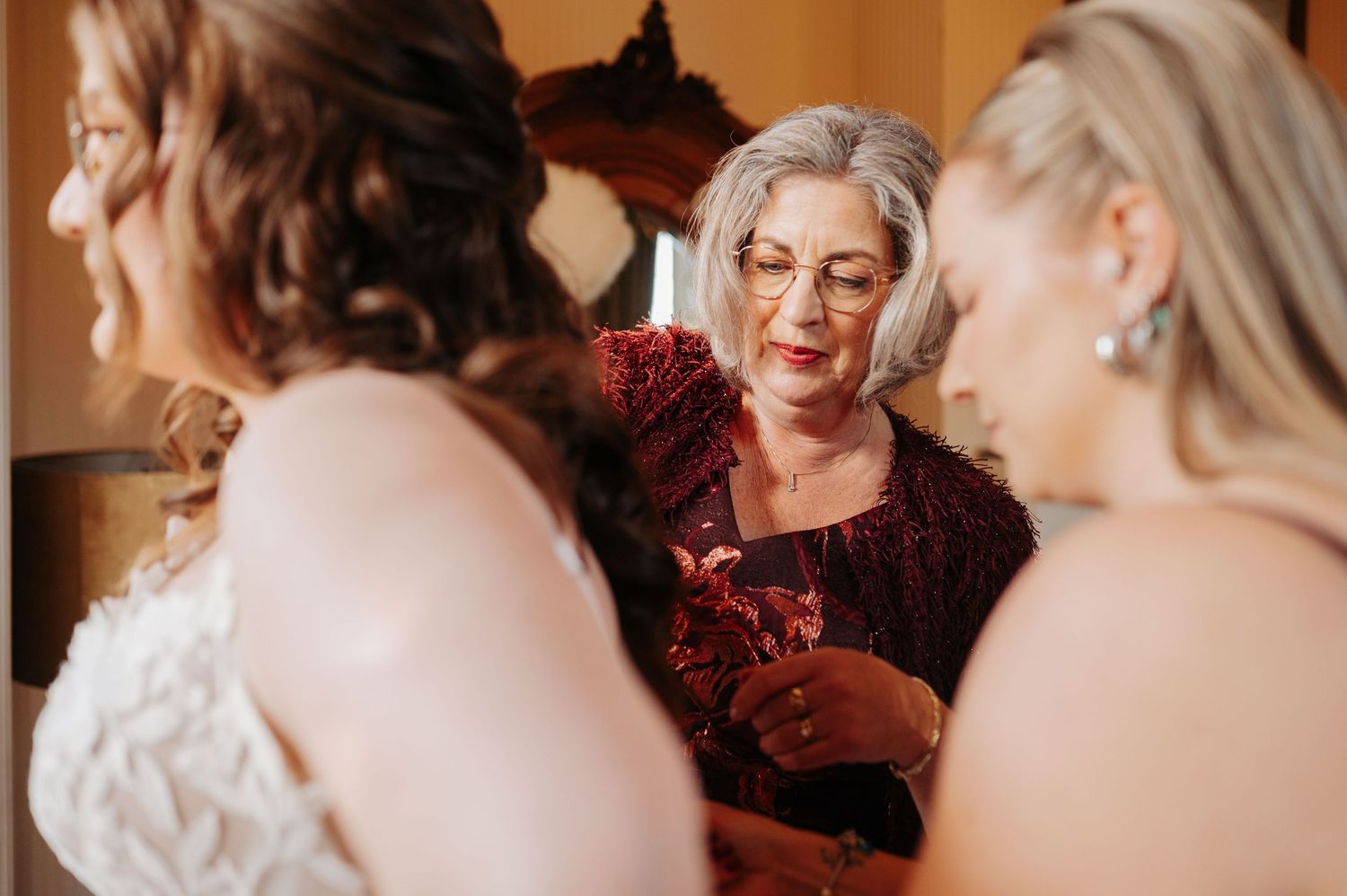 Smiling older woman in a burgundy lace top helps a bride in a white lace gown get ready while another woman looks on.