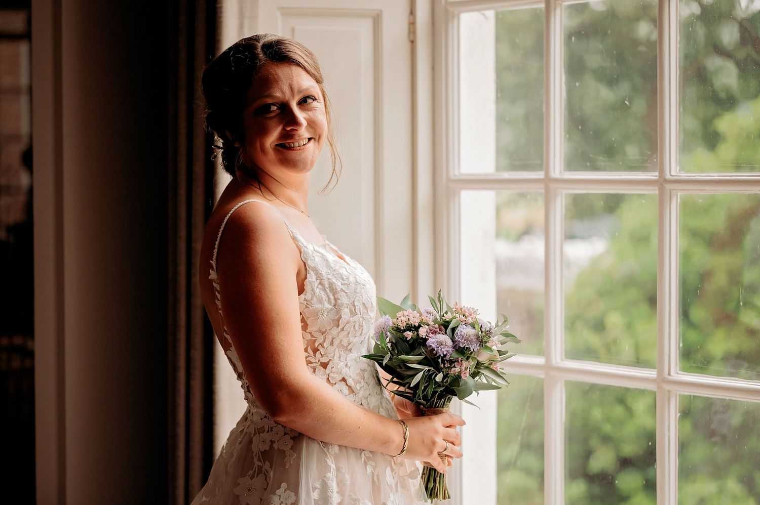 Bride in lace wedding dress holding purple bouquet standing by large window in elegant indoor setting.