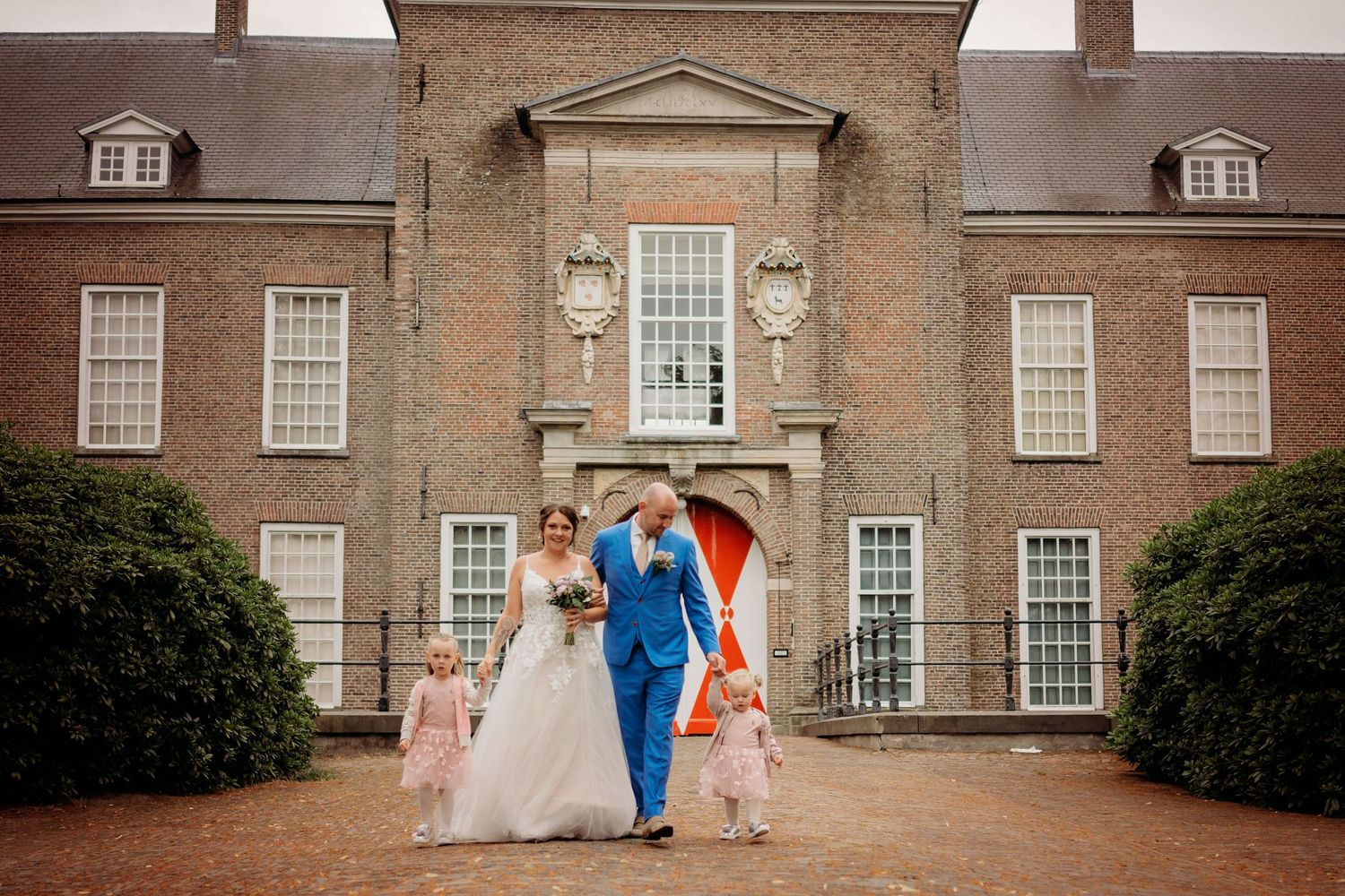 Bride and groom walk hand in hand with flower girls outside a historic brick mansion on their wedding day.