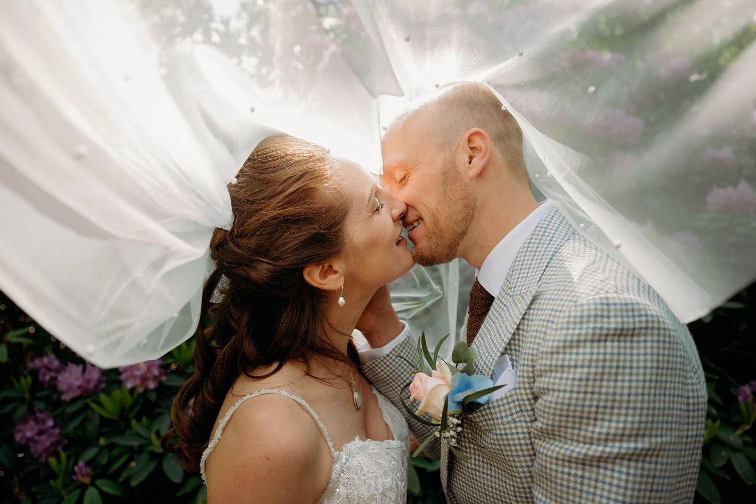 Bride and groom sharing a romantic kiss under a flowing white veil in a garden.