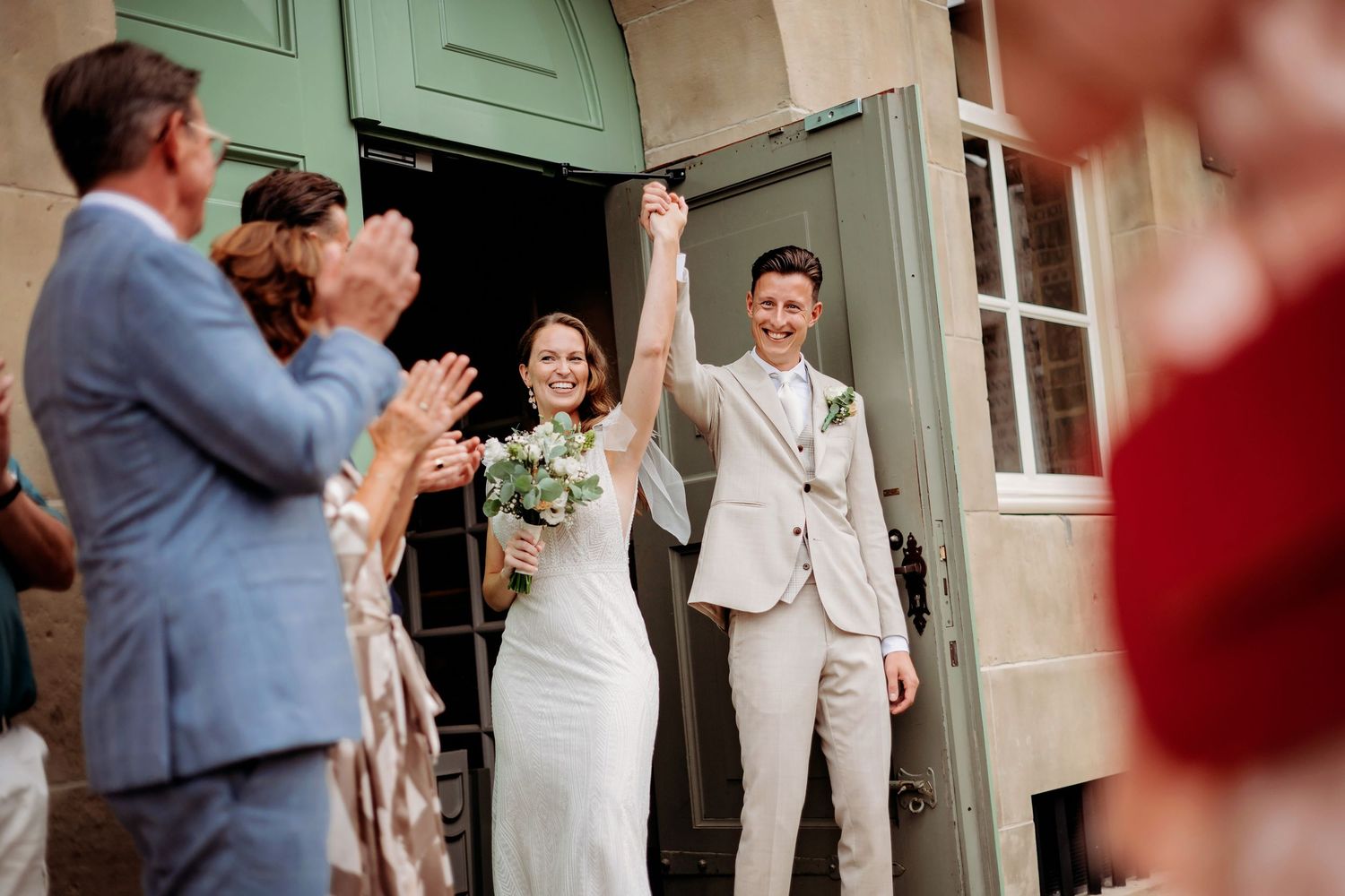 Joyful newlyweds exit church doors raising hands in celebration while guests applaud, bride holding white bouquet.