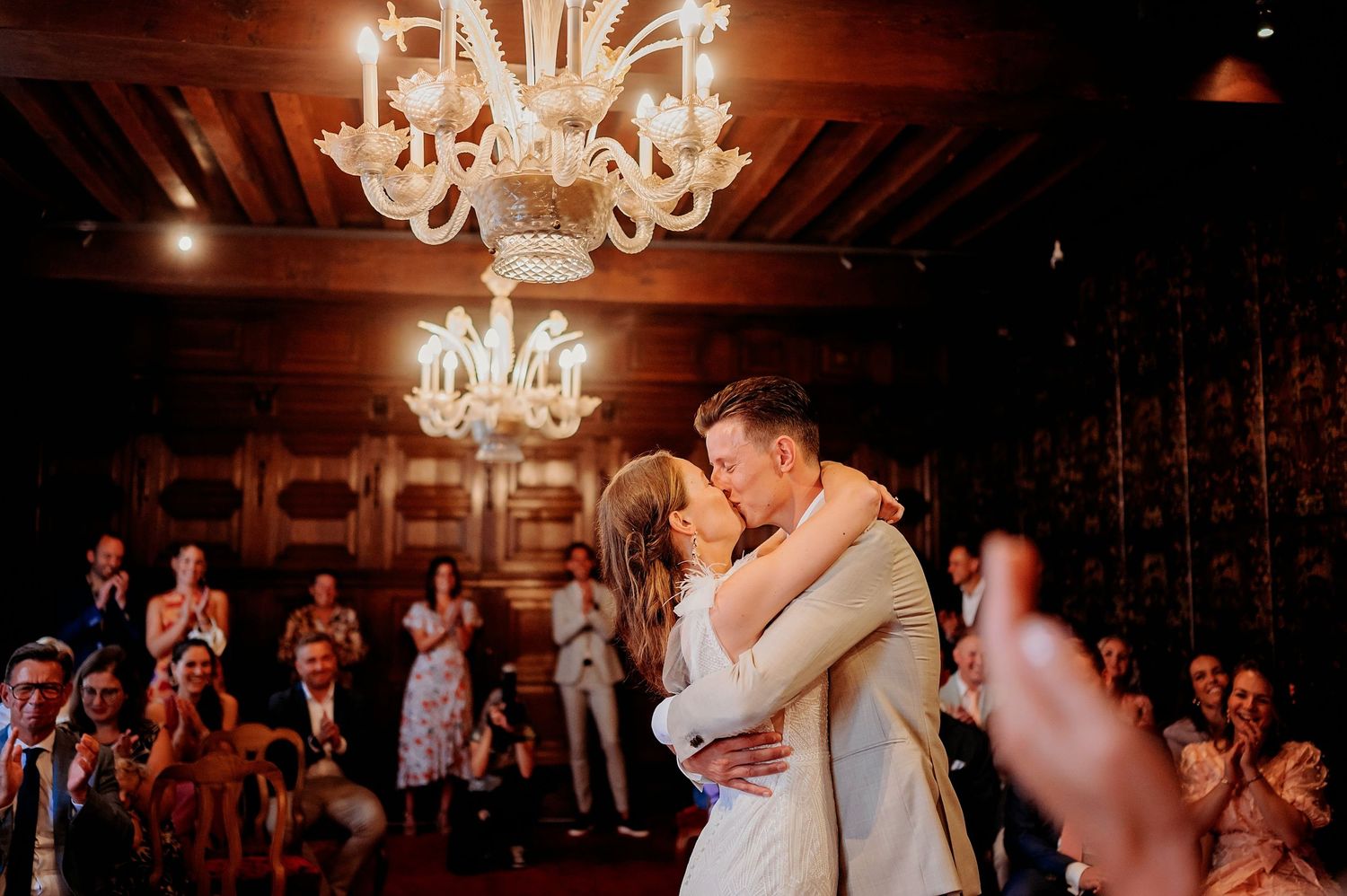 Bride and groom share first dance kiss under elegant chandelier surrounded by applauding wedding guests in warmly lit venue.