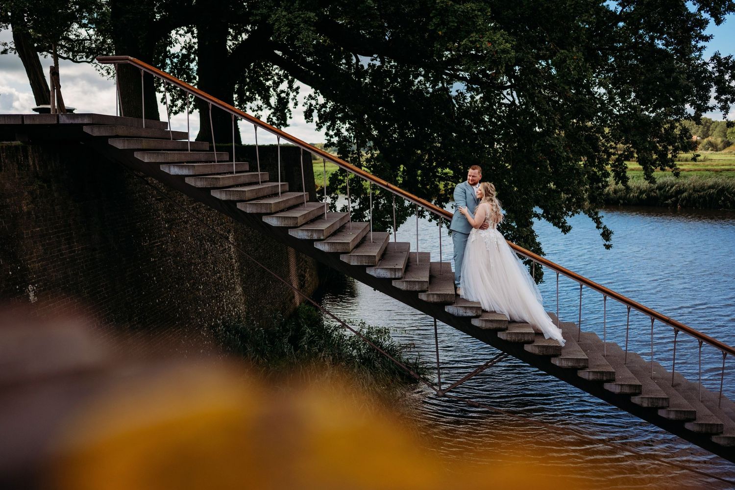 Bride and groom embracing on stone bridge stairs by a river, surrounded by lush green trees on their wedding day.