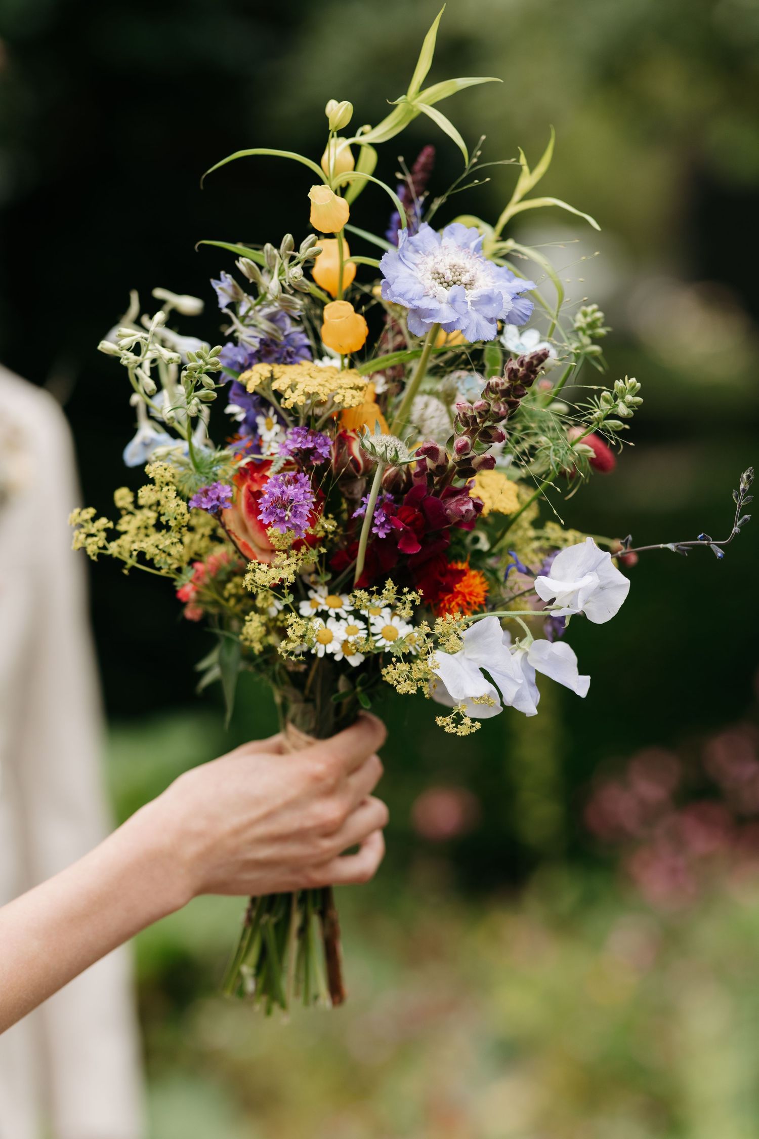 Hand holding a vibrant wildflower wedding bouquet with blue, yellow, red, and white blooms against a garden backdrop.