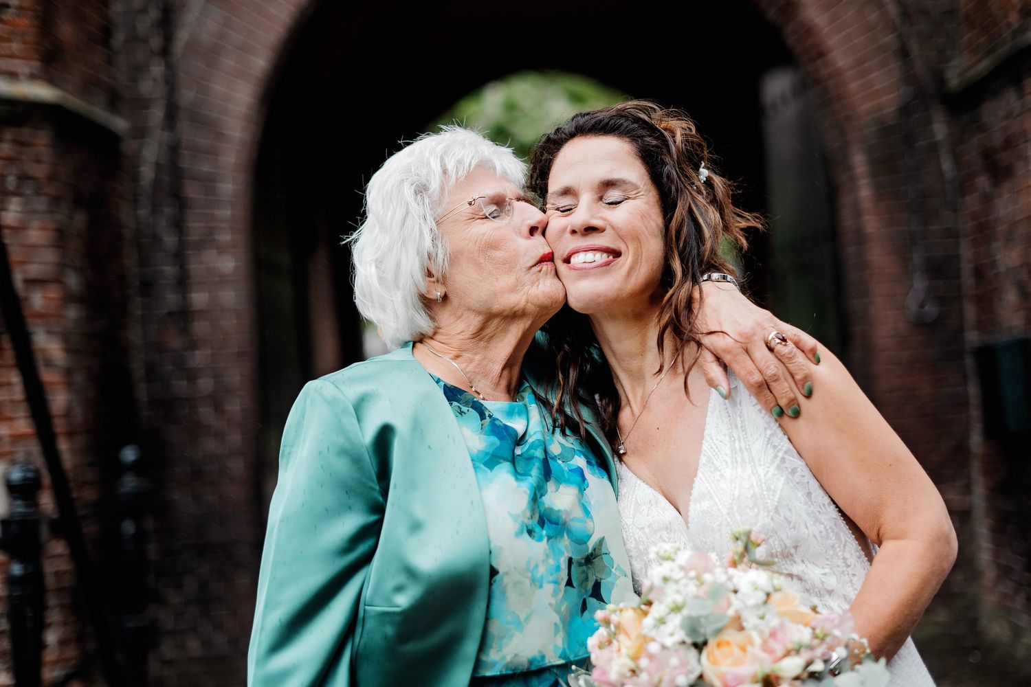 Teder moment tussen twee generaties met pastelboeket en bakstenen boog, bruidsfotografie Vught, Brabant.