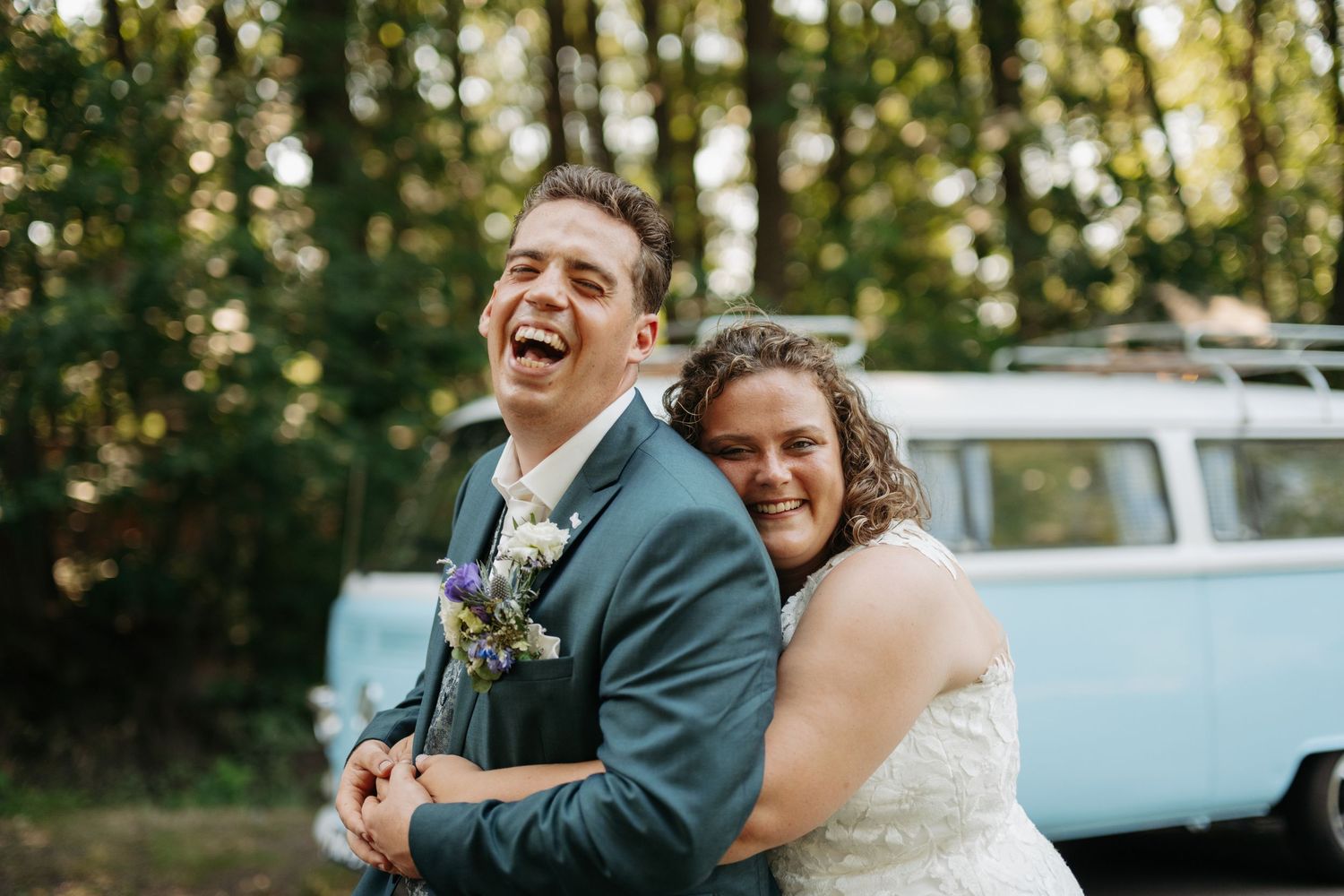 Laughing bride and groom embracing outdoors in front of a vintage blue VW bus at their wedding.