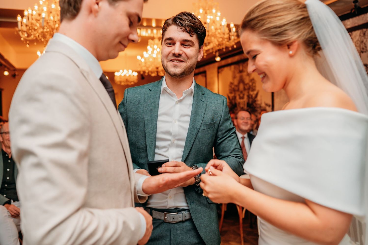 Bride and groom exchanging rings during wedding ceremony with smiling officiant in teal suit indoors.