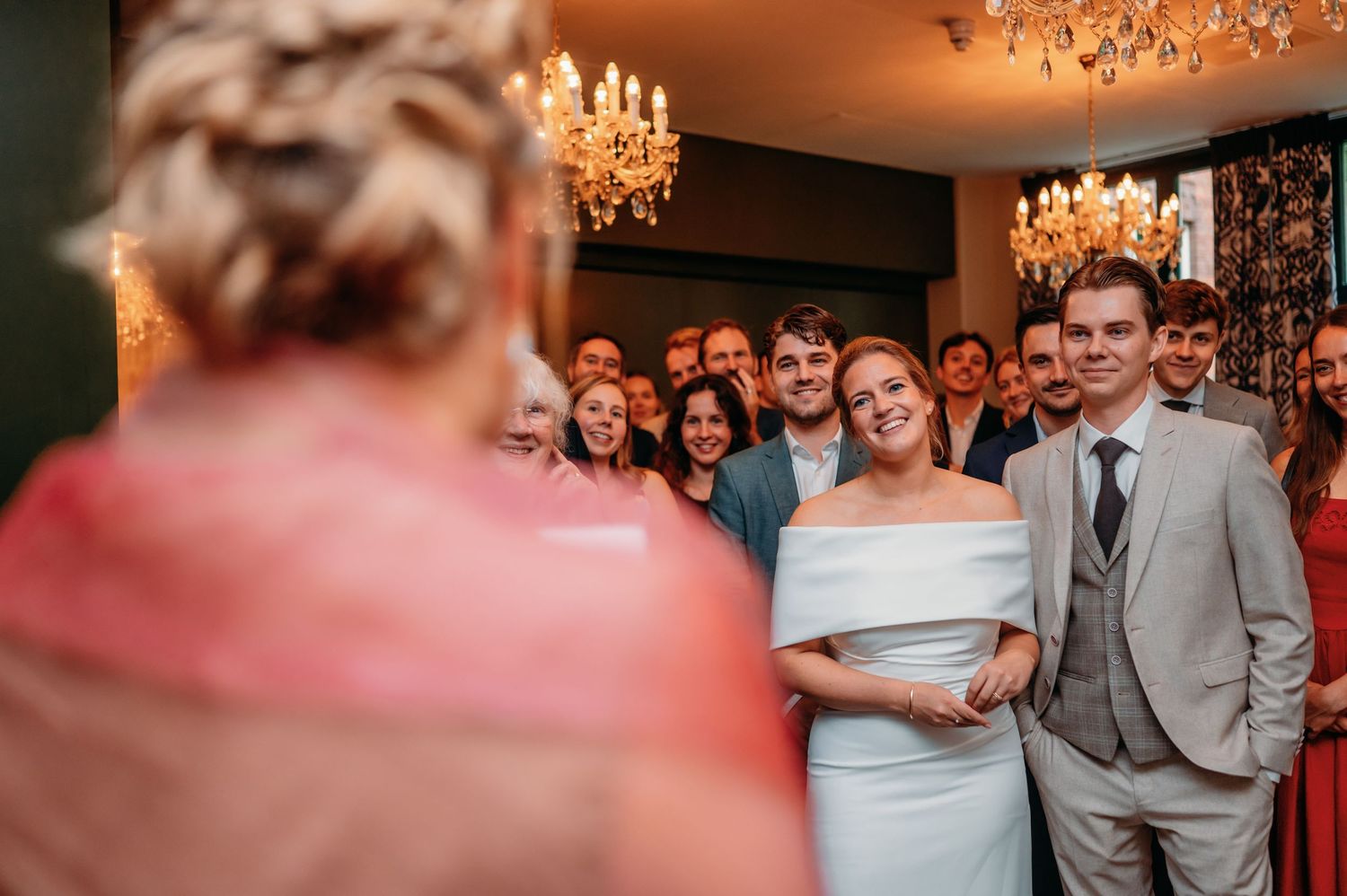 Bride and groom smiling at wedding guests in elegant chandelier-lit reception room.