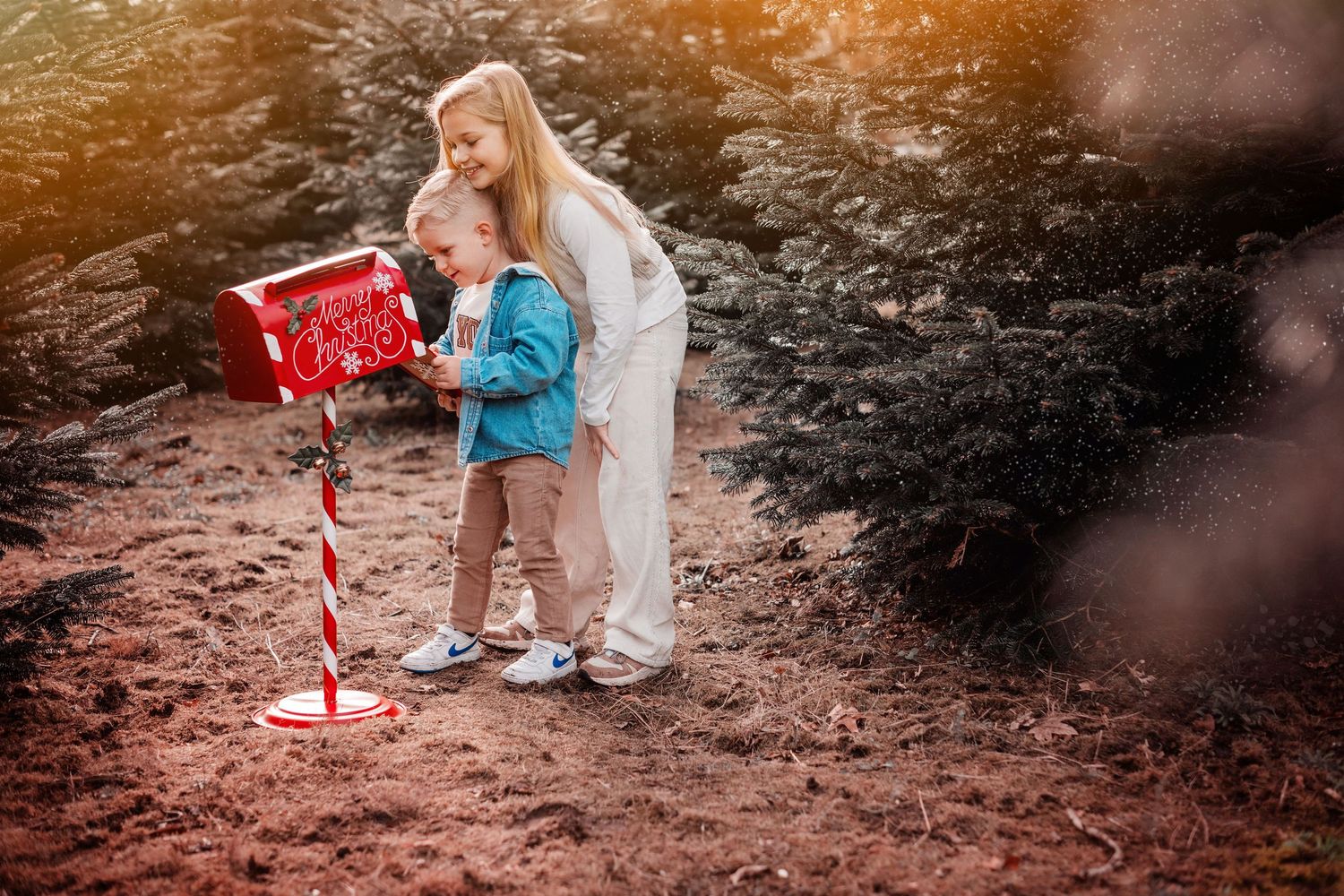 A mother and child mailing a letter to Santa at a festive red mailbox surrounded by Christmas trees.