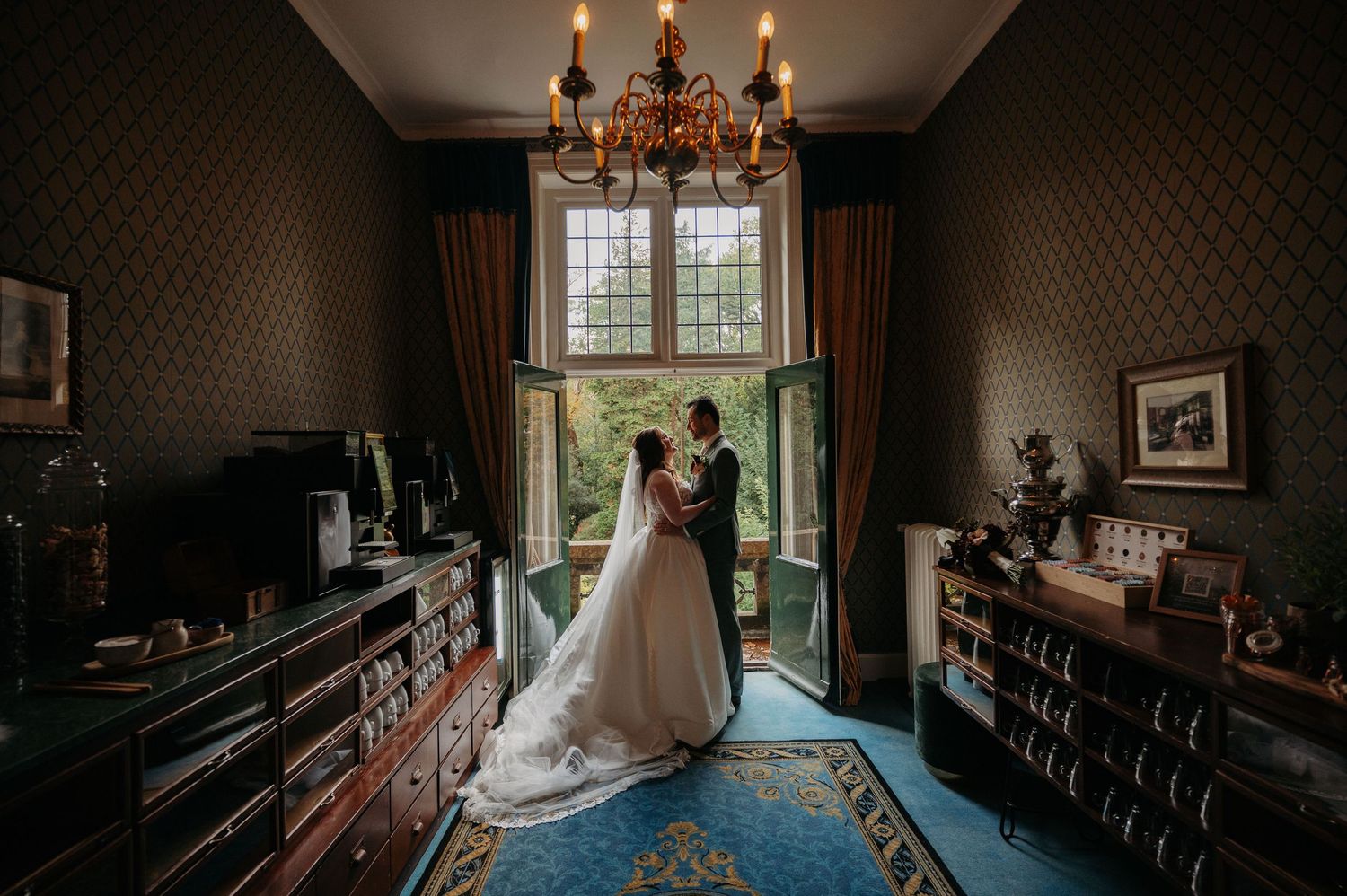 Bride in a flowing white gown and groom embrace by a tall window in an elegant vintage room with a chandelier and ornate rug.