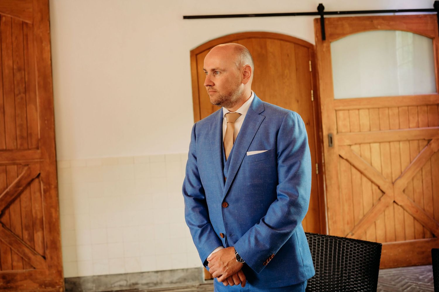 Dapper groom in blue suit with tan tie stands confidently indoors before wedding ceremony.