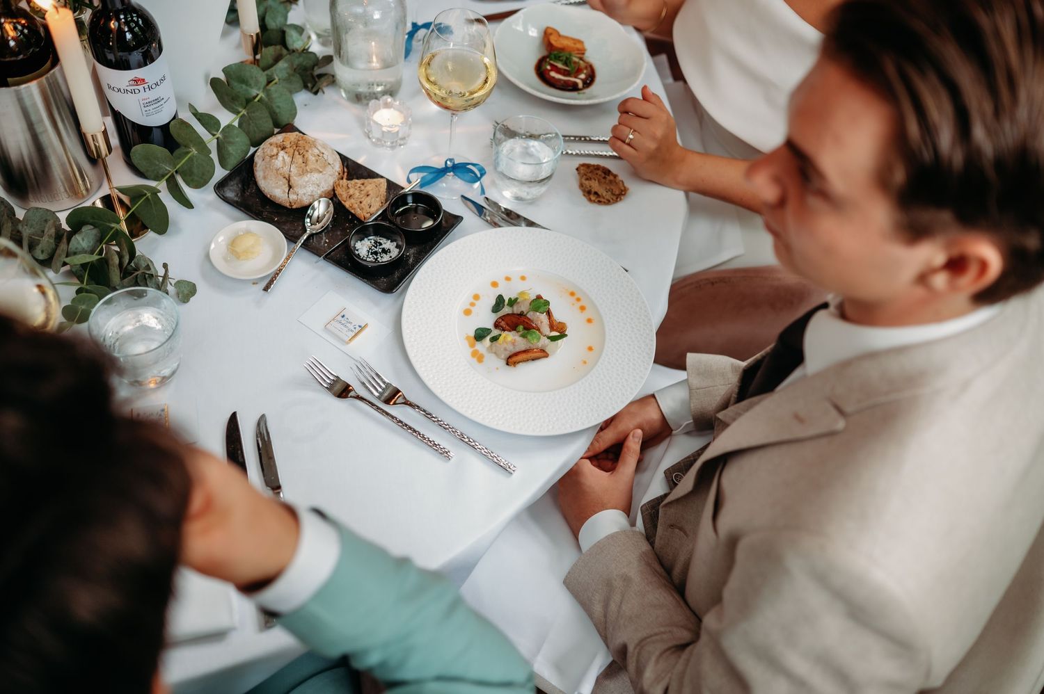Overhead view of wedding guest in tan suit at elegant reception table with plated gourmet dish and floral centerpiece.