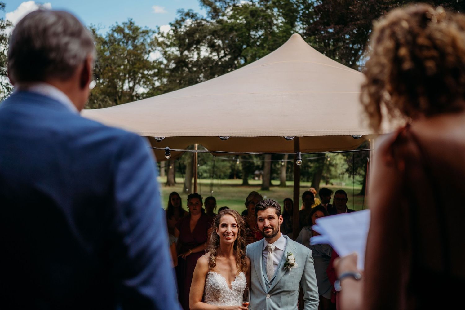 Couple exchanging vows at outdoor ceremony under beige tent with guests watching.