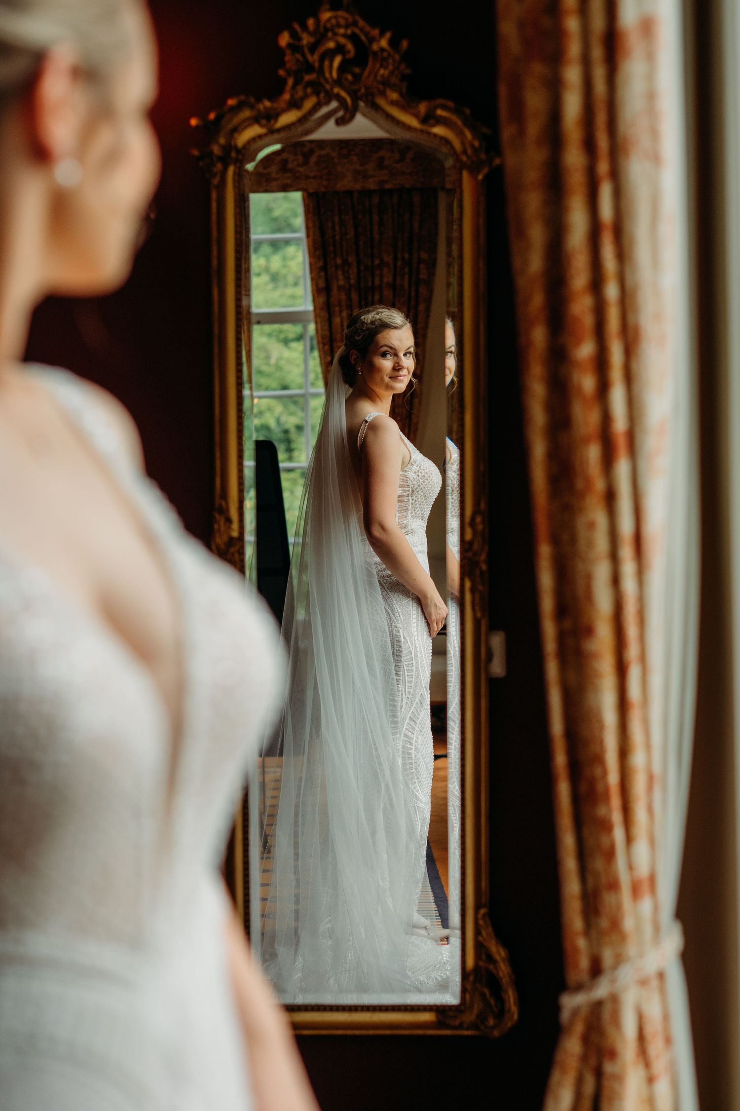 Elegante bruid poseert in witte jurk voor gouden spiegel in klassieke interieursetting, bruidsfotografie Brabant