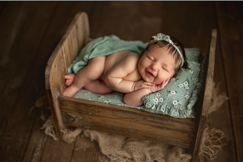 Smiling newborn baby sleeping peacefully in a rustic wooden bed with mint green floral bedding and headband.