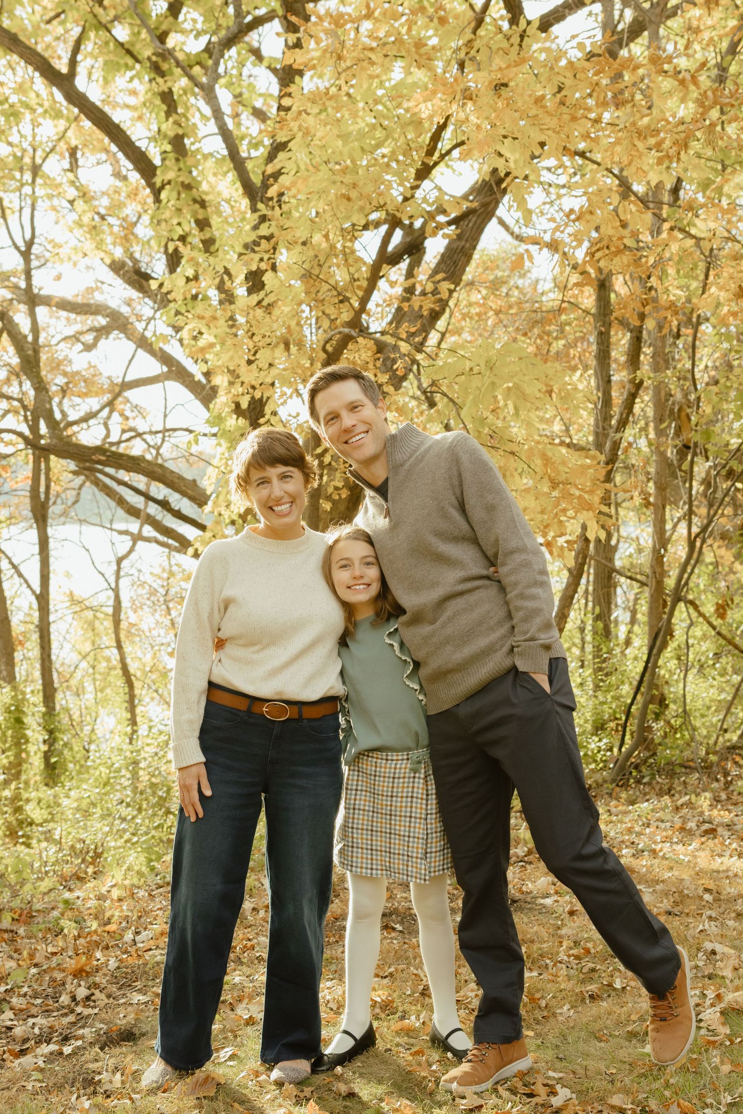 Family of three smiling together in autumn forest with golden leaves, wearing casual sweaters and jeans.
