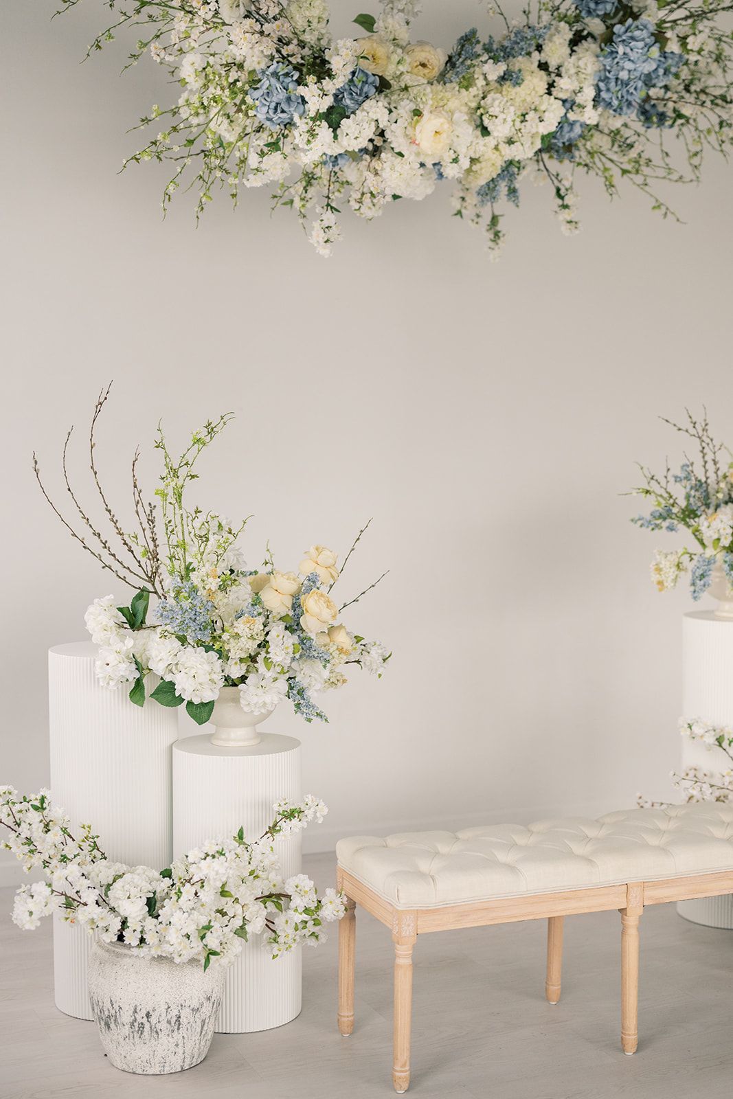 Elegant wedding decor with white and blue hydrangea floral arrangements on pedestals, a tufted bench, and overhead flower installation.
