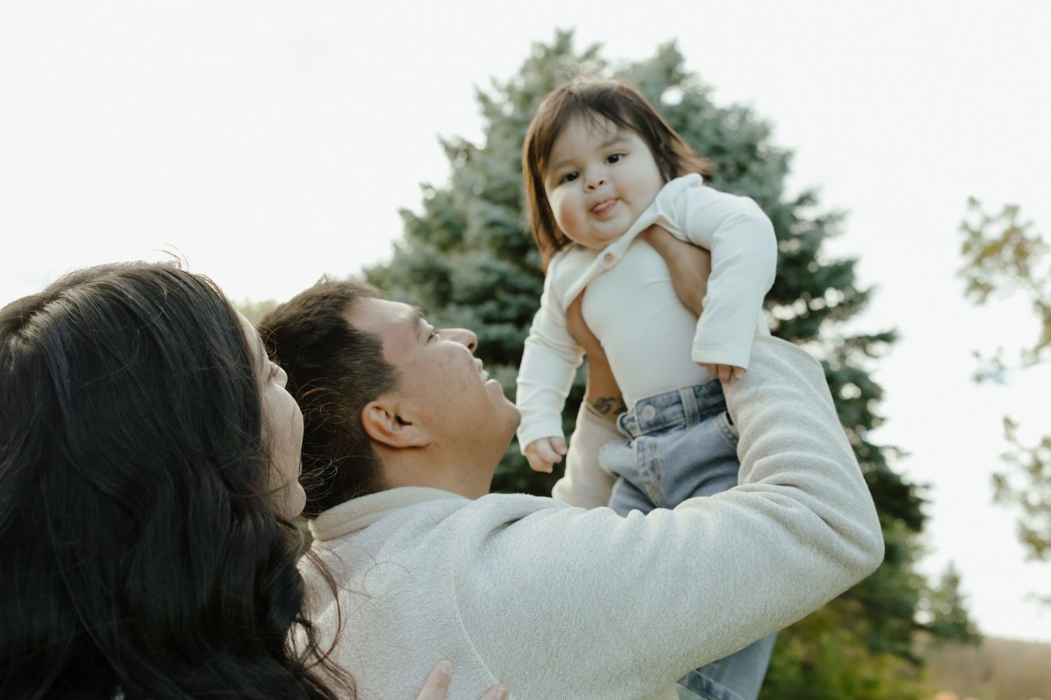 Parents joyfully lifting their baby girl outdoors near a green tree on a bright day.