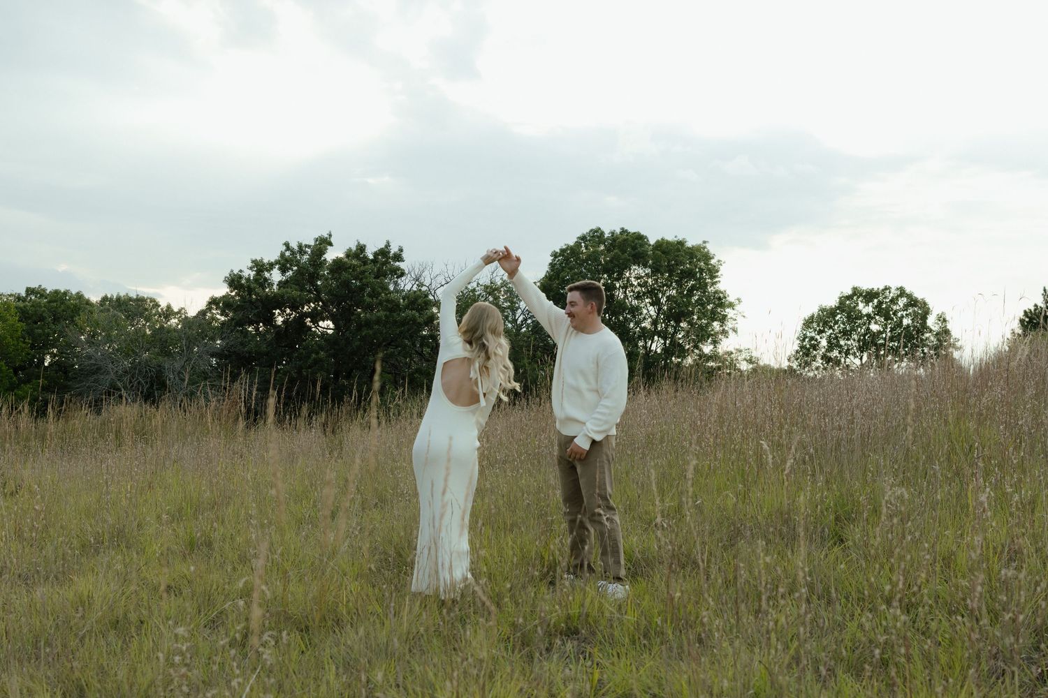 A couple in white attire stands together in a grassy meadow on an overcast day.