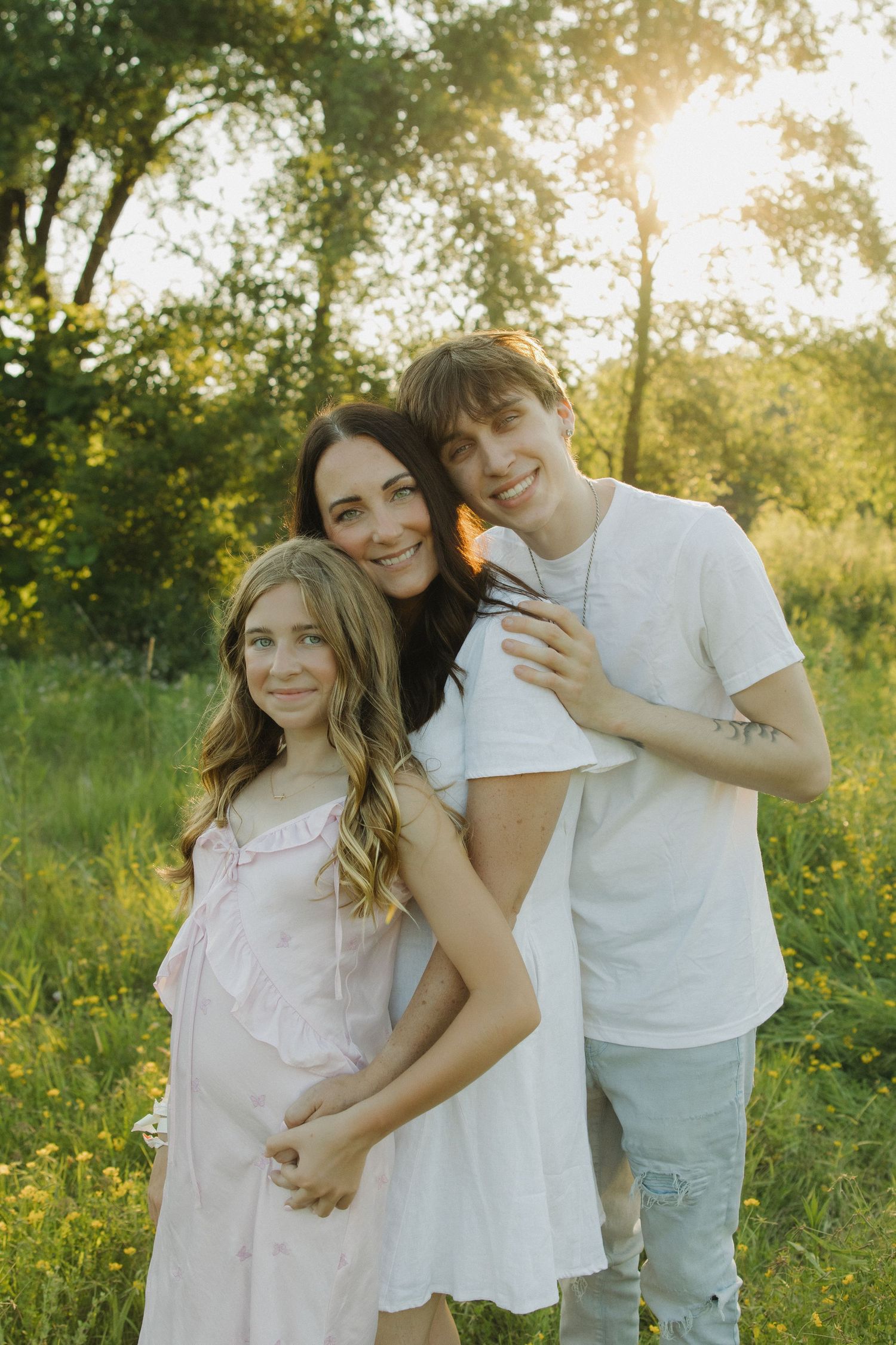 A woman and two teenagers pose together in a sunlit green meadow, smiling warmly in a family portrait.