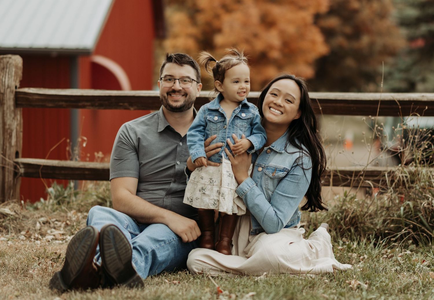 A couple sits together on grass near a rustic red barn during an autumn engagement photo session.
