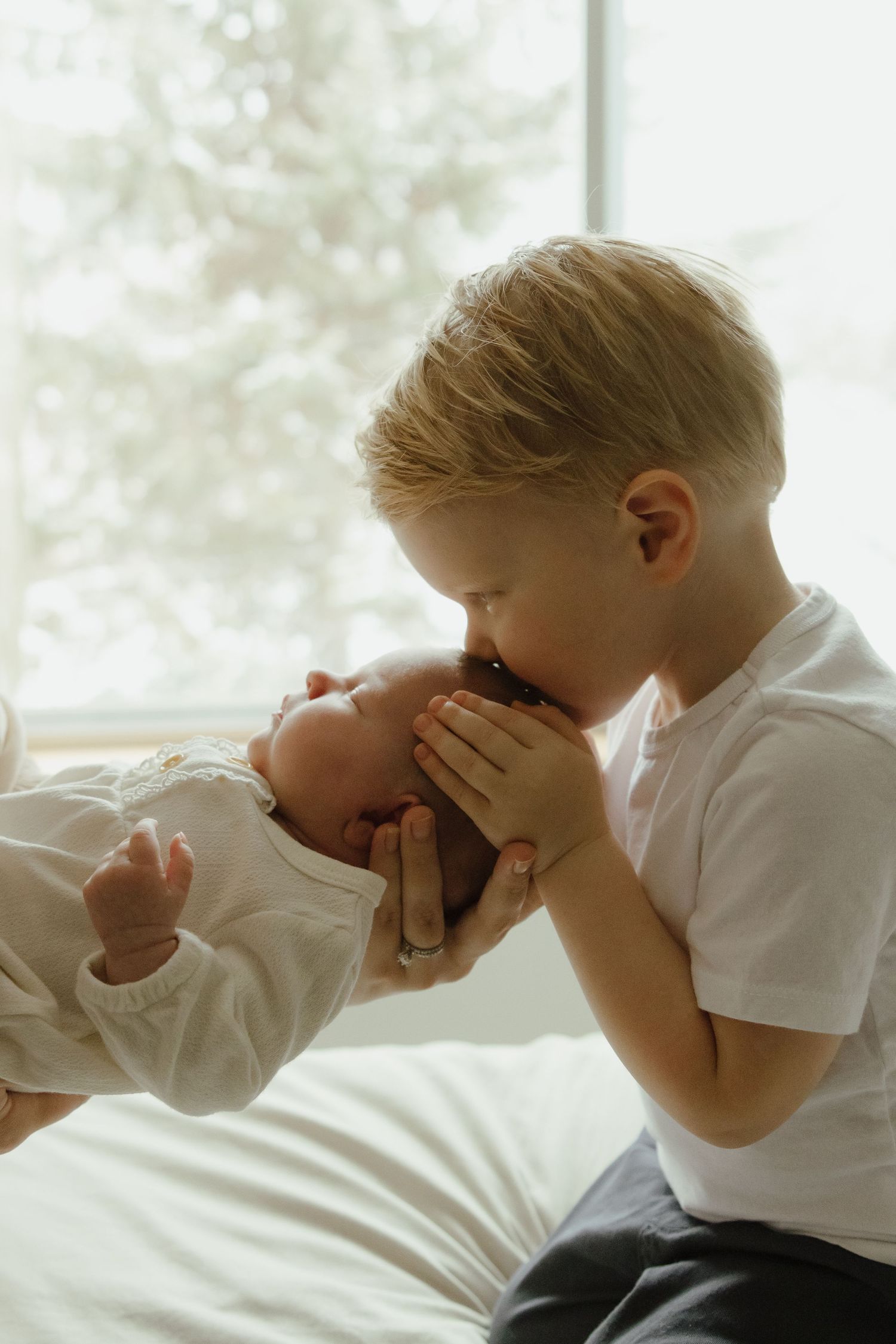A young blonde boy gently kisses the head of a newborn baby, cradling it lovingly near a bright window.
