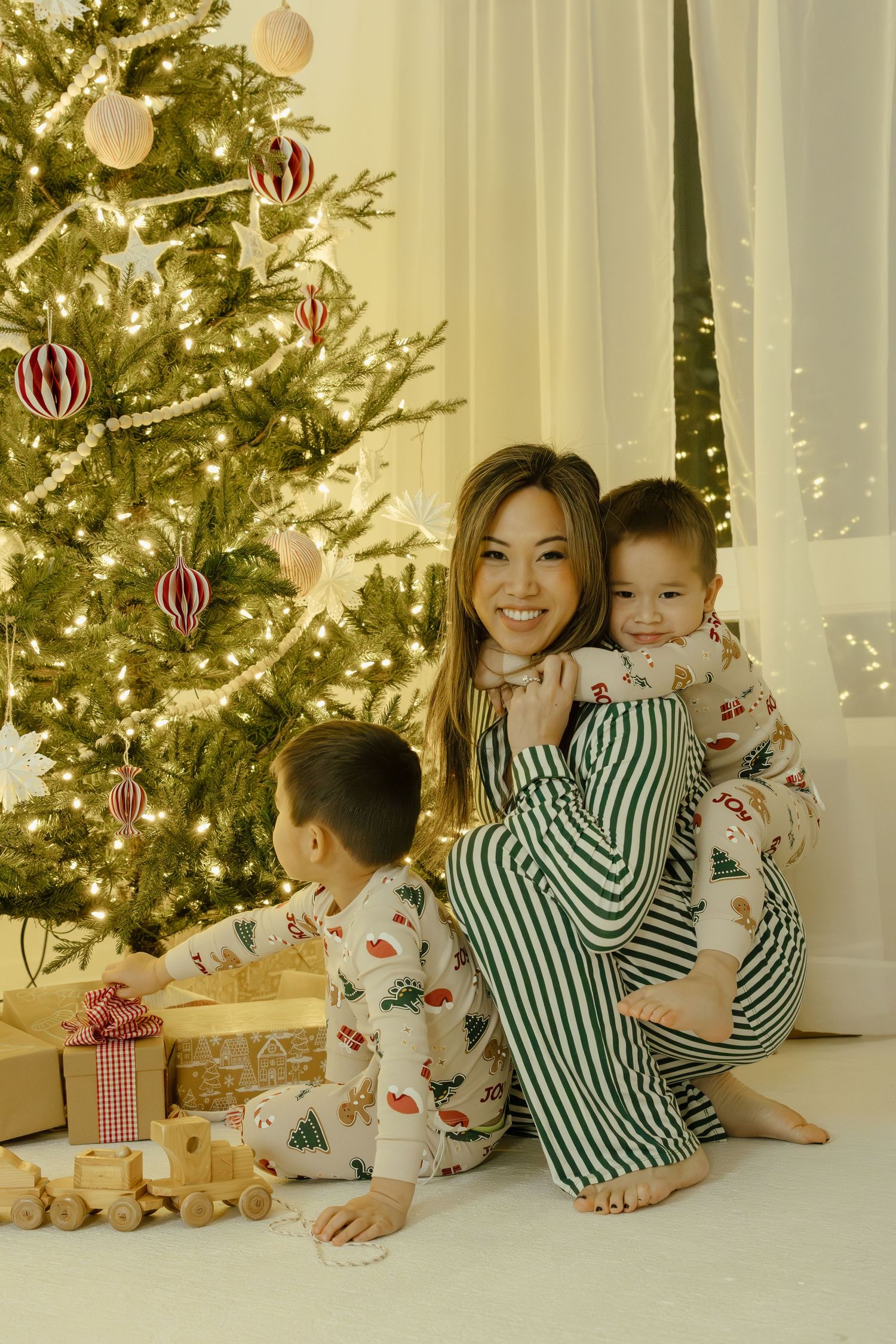 Mom and two children in matching Christmas pajamas sitting by a decorated tree with gifts on a white floor.