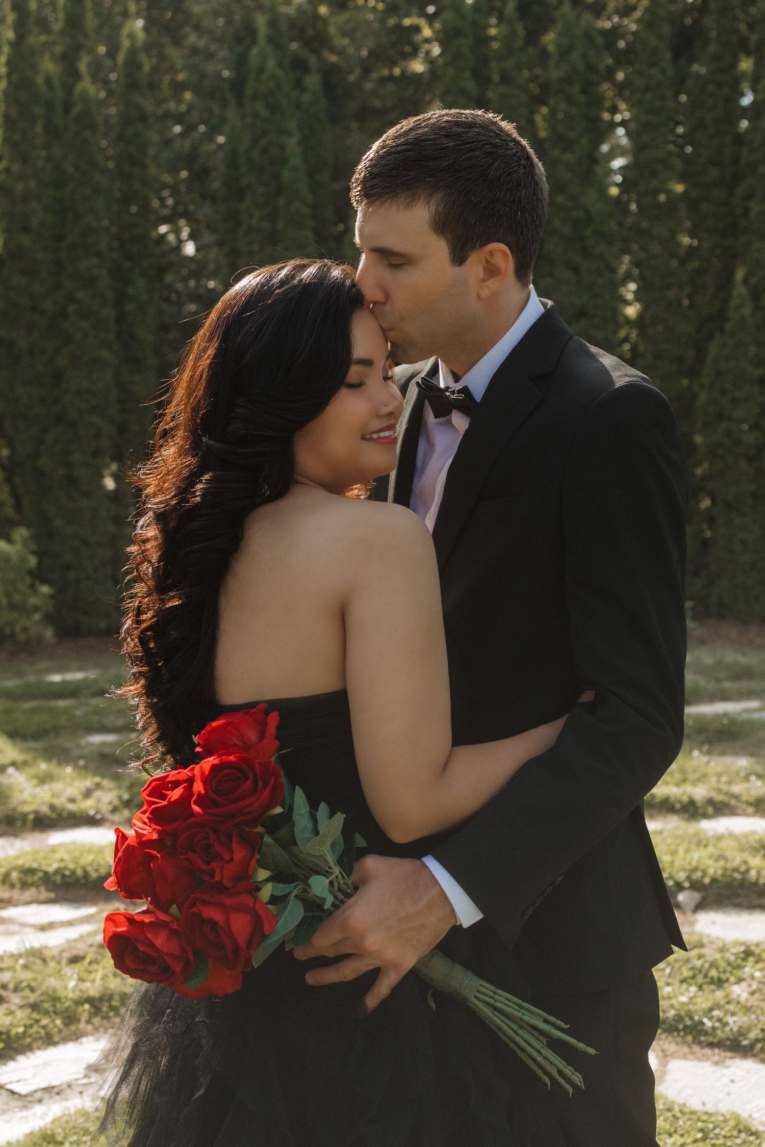 A couple in formal attire shares an intimate moment with red roses in a garden.
