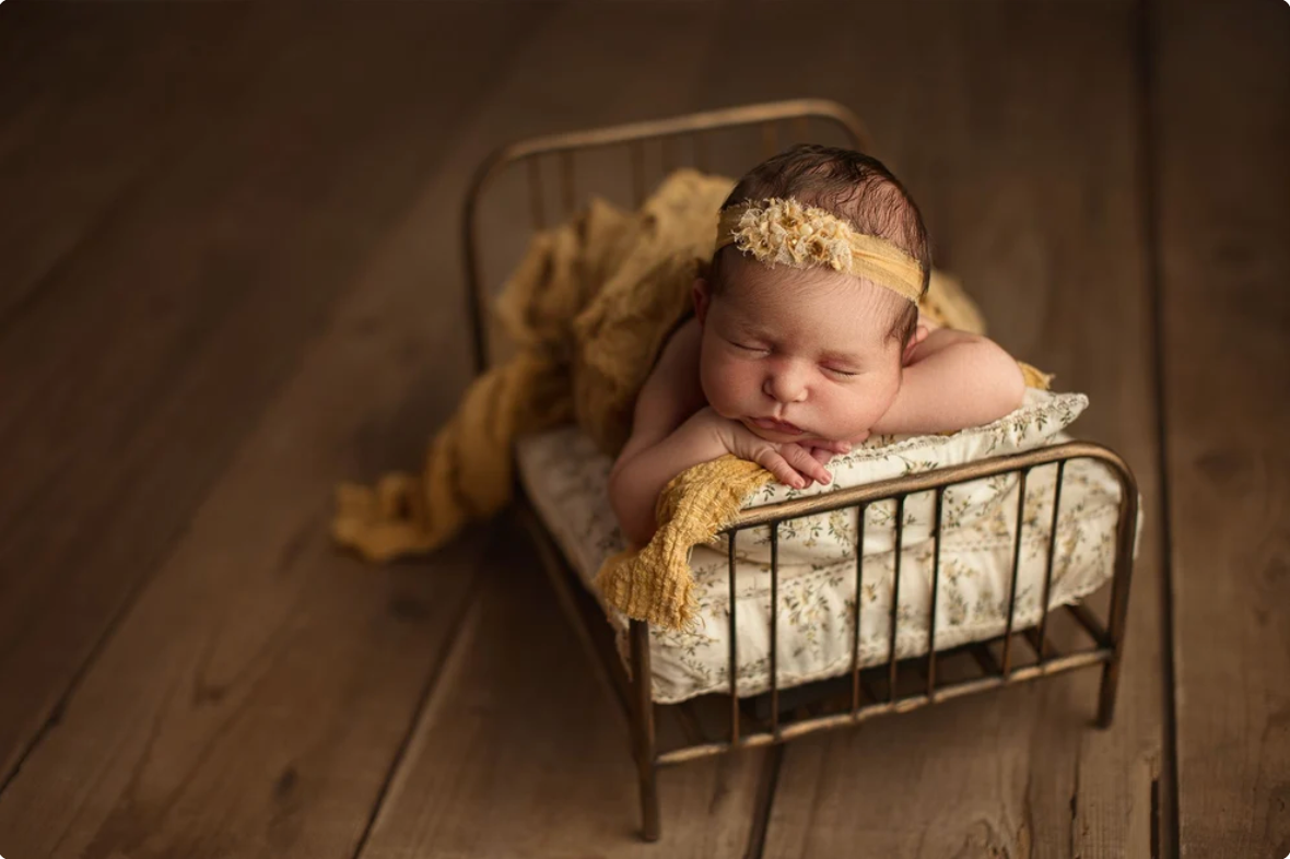 Newborn baby sleeping peacefully in a miniature gold bed with yellow lace blanket and floral headband on wood floor.