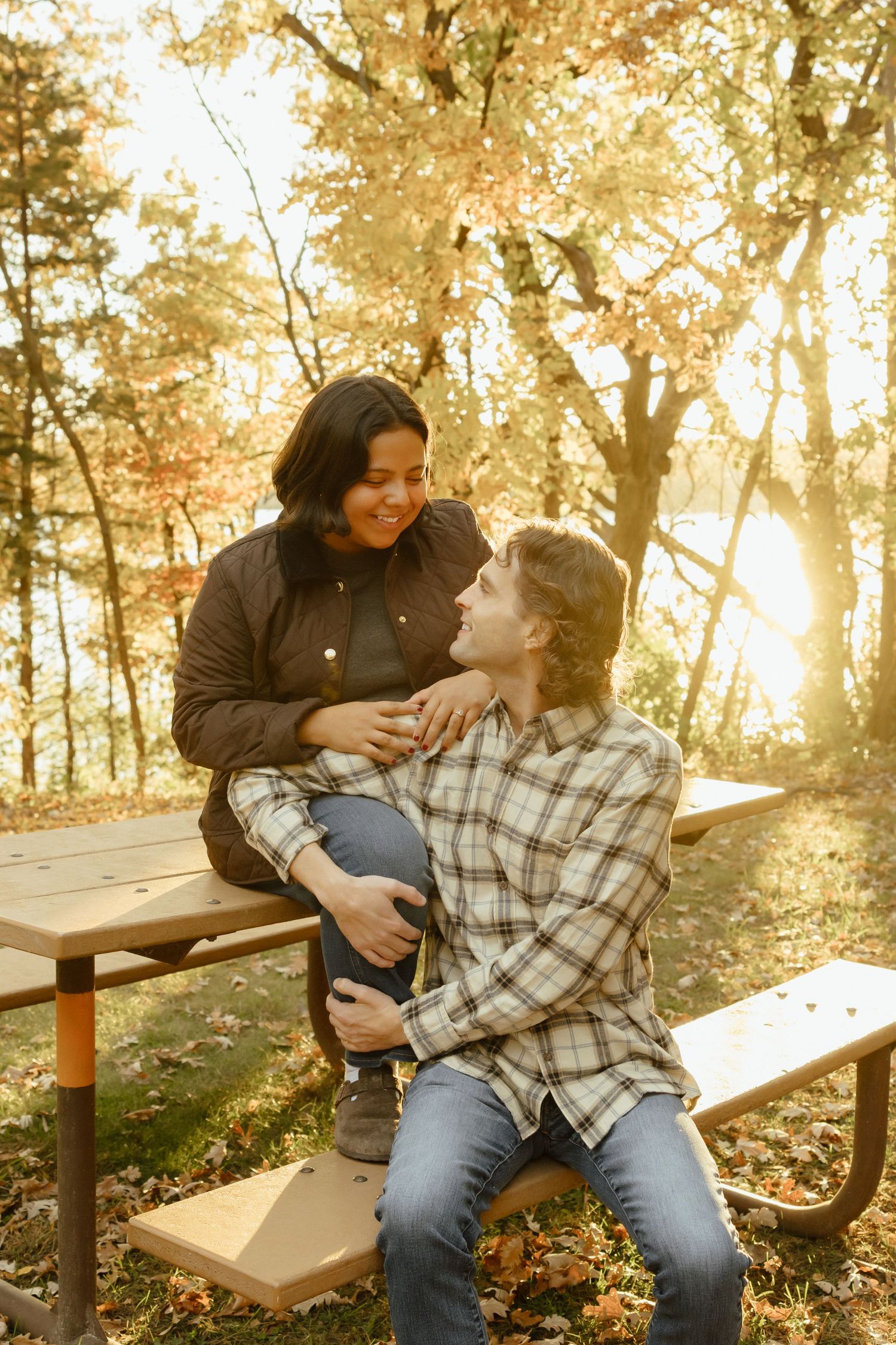 A couple shares a tender moment sitting on a wooden bench in a sunlit autumn forest.