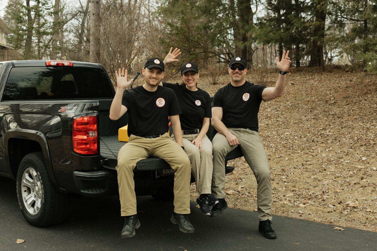 Three people in matching black shirts and khaki pants sit on the tailgate of a black pickup truck while waving at the camera.