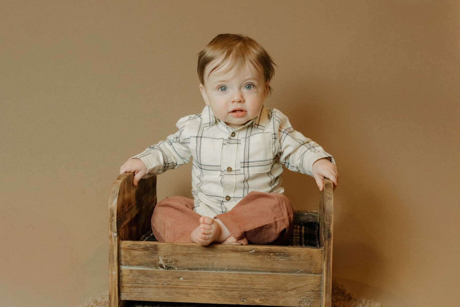 Small child in plaid shirt and coral pants sits in rustic wooden crate against neutral background.