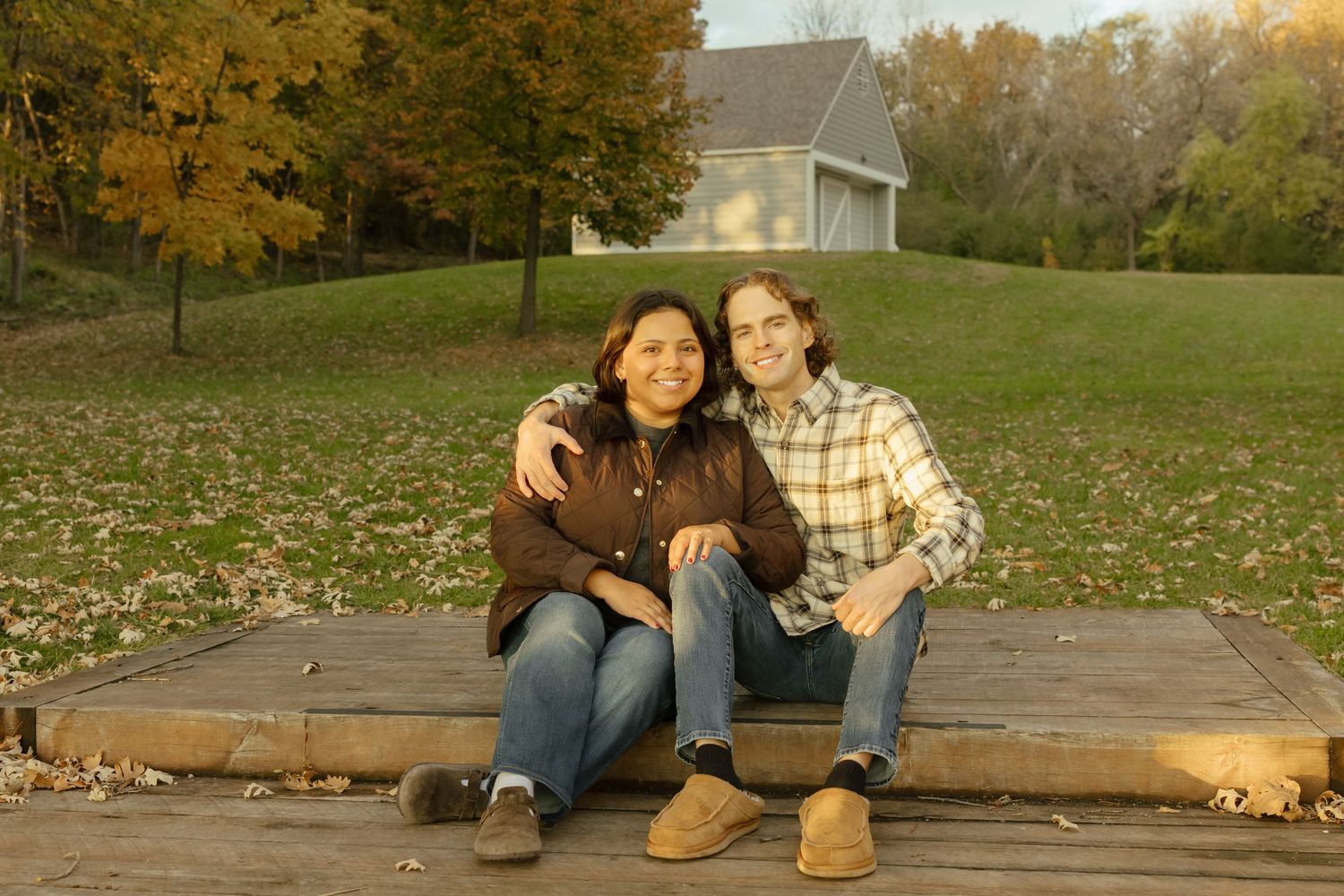 Couple sitting together on wooden deck outdoors, smiling, surrounded by autumn foliage and a house in background.