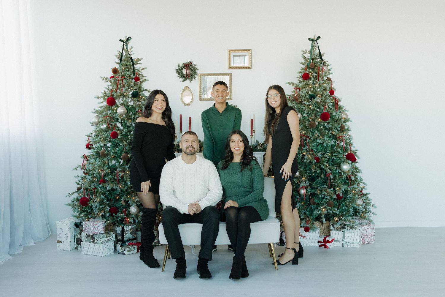 Family of five posing for a Christmas photo with decorated trees, gifts, and holiday decor in a bright studio.