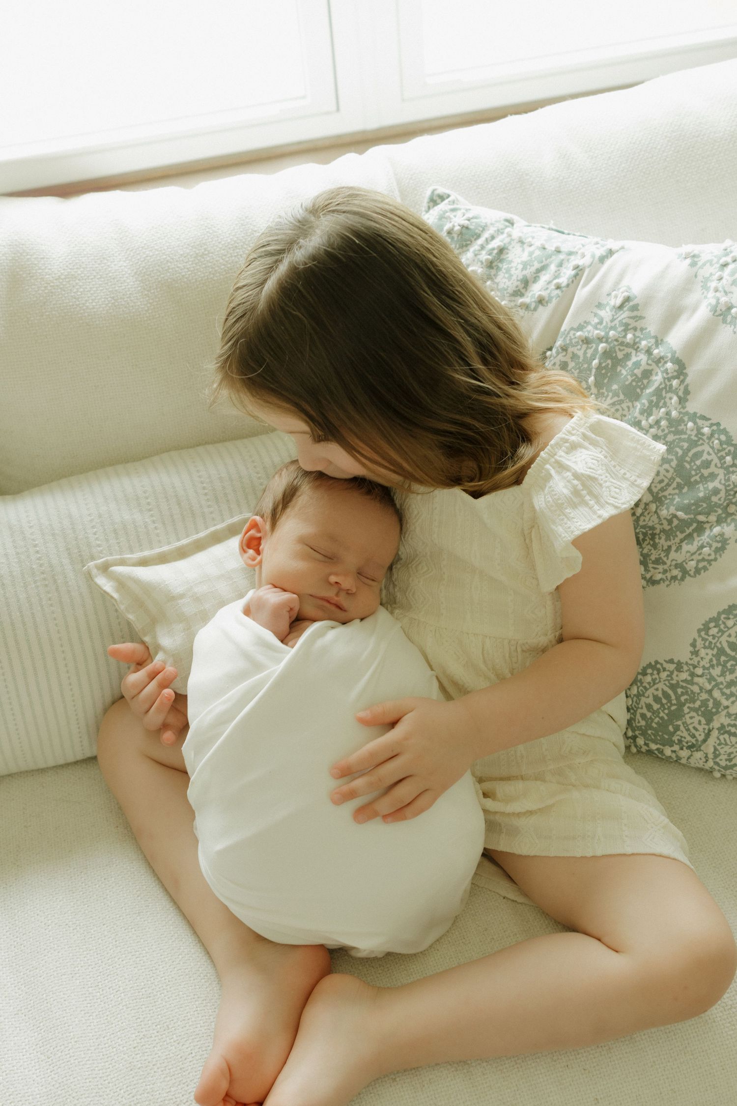 Young girl sits on cream sofa gently cradling sleeping swaddled newborn baby, sharing a tender sibling moment.