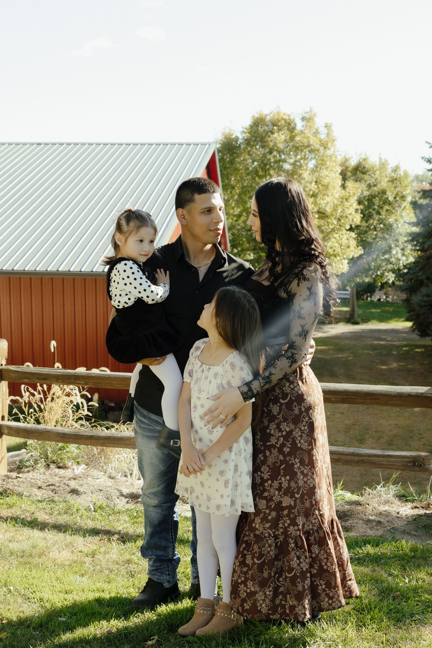 A family of four poses outdoors on a farm, with parents holding young daughters near a red barn on a sunny day.