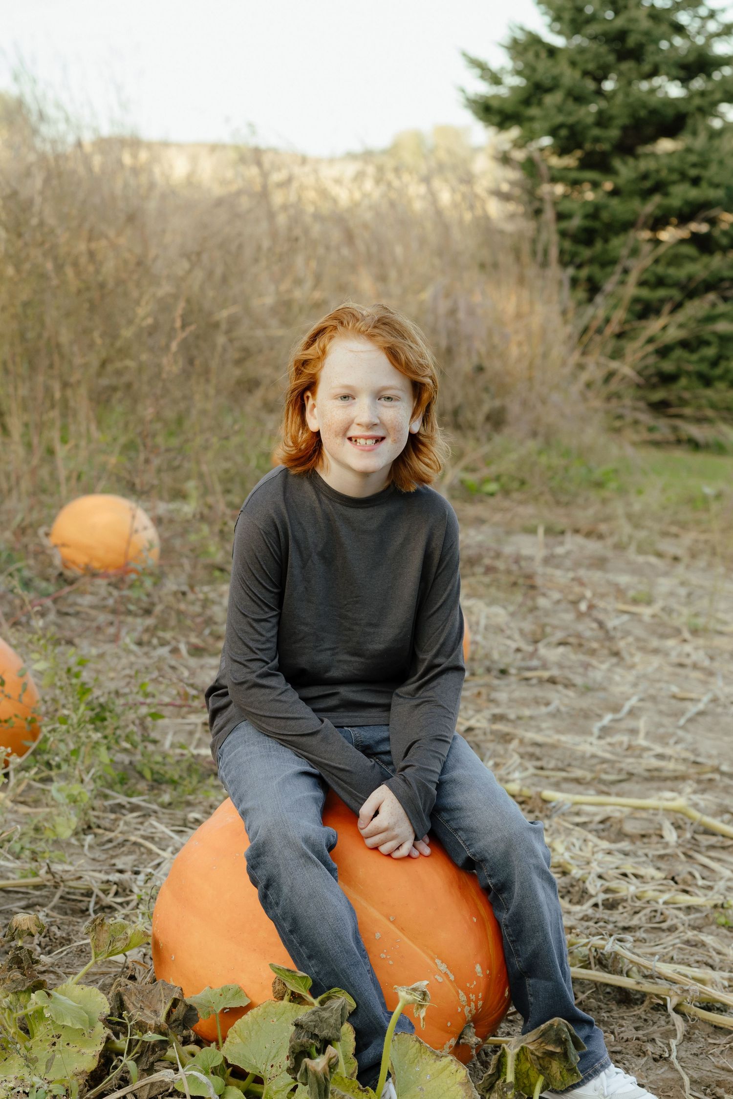Red-haired child sitting on a large orange pumpkin in a fall pumpkin patch, smiling outdoors.
