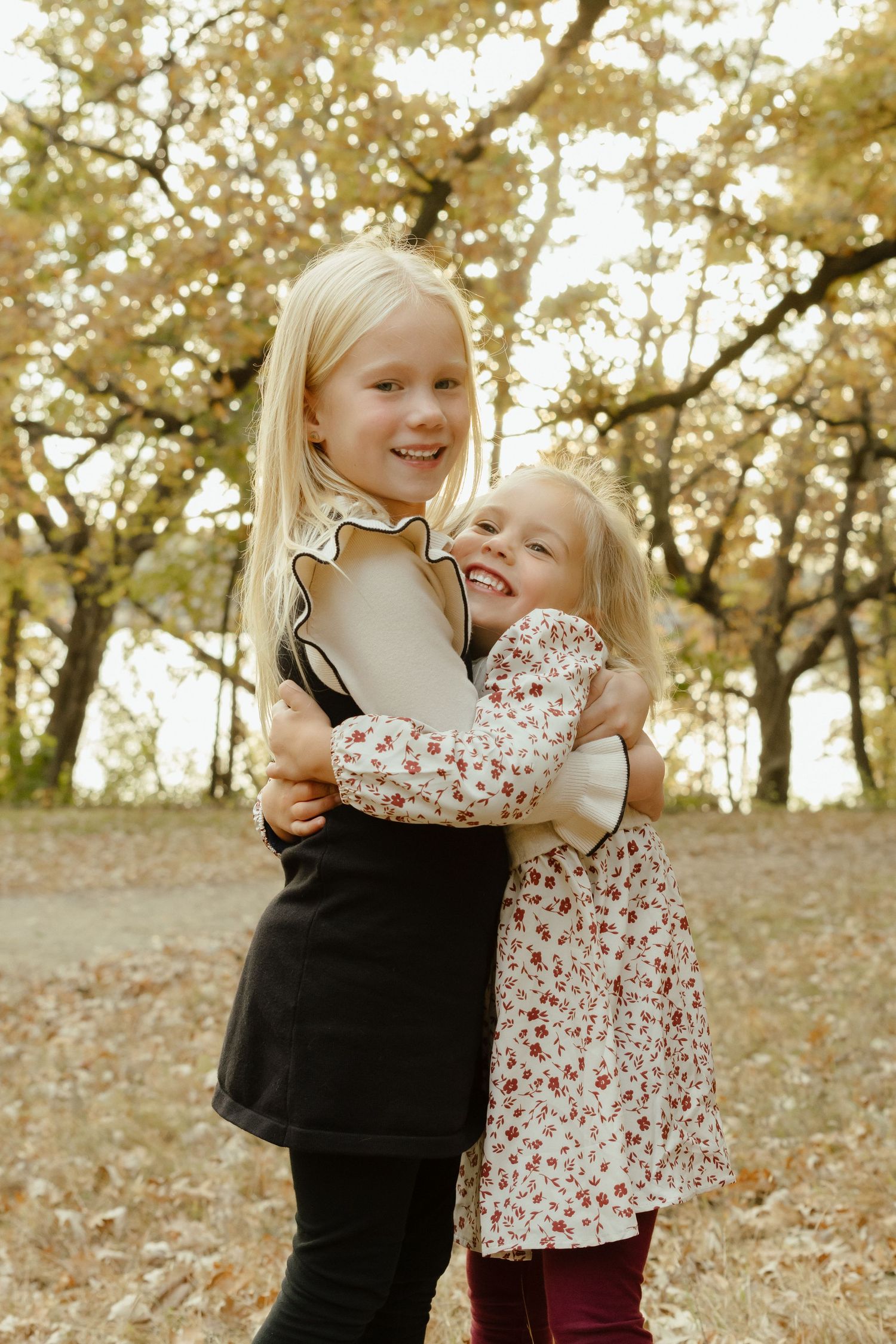 Two blonde sisters hugging and smiling in an autumn park with golden trees and fallen leaves.